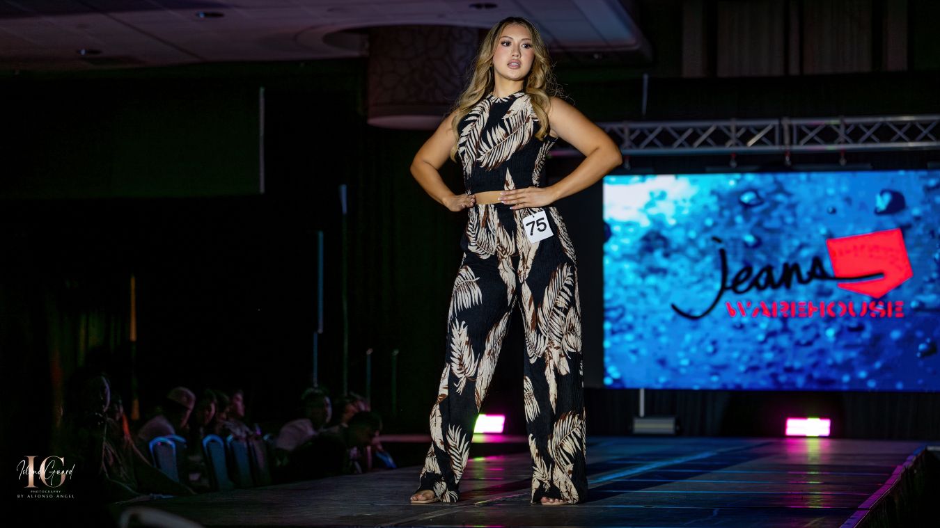Model showcases black and white patterned jumpsuit on fashion runway with blue backdrop.