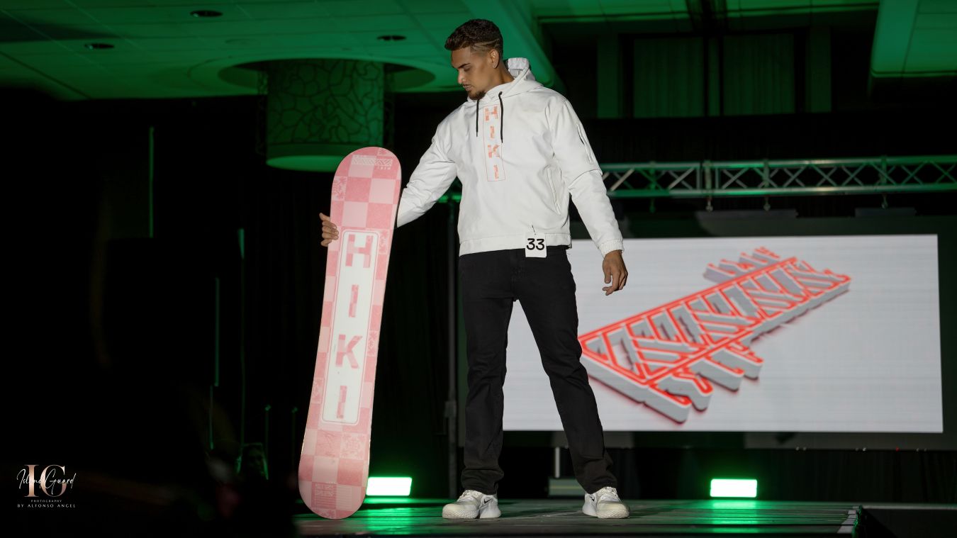 Model in dark attire poses dramatically on spotlit fashion catwalk.