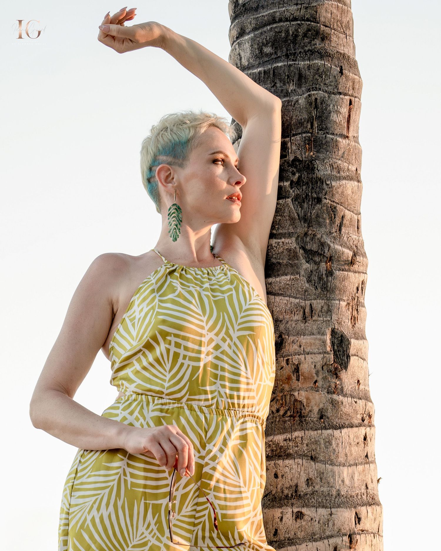 Person in yellow and white palm-print dress poses gracefully against tropical palm trees at sunset.