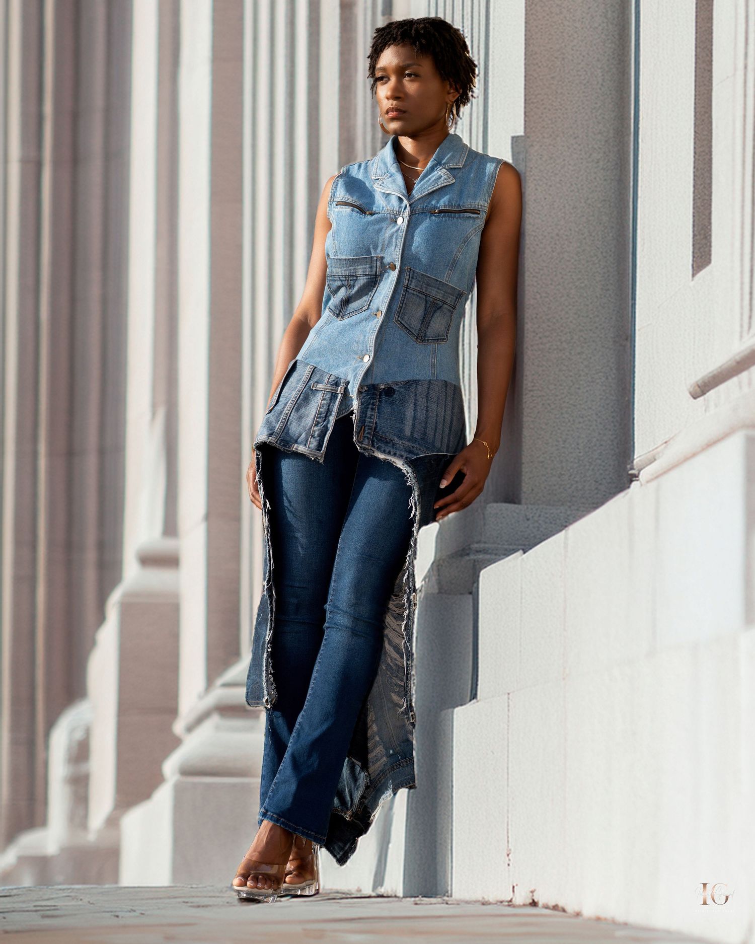 Model wearing denim vest and jeans poses against white columned backdrop.