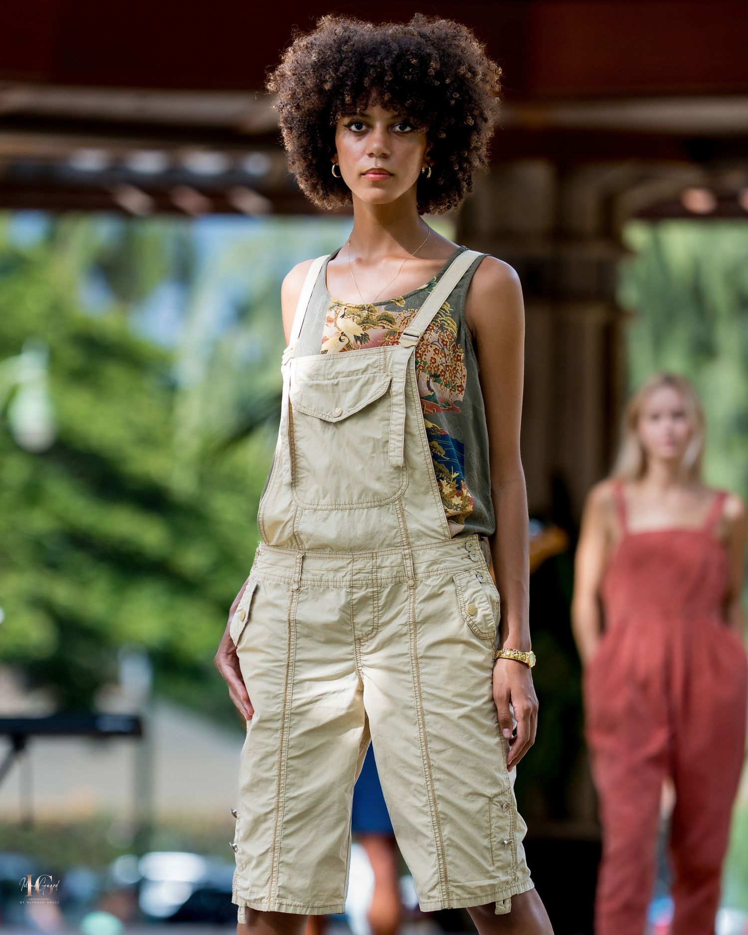 Person wearing beige cargo overalls shorts and natural curly hair poses on outdoor walkway with blurred figure in background.