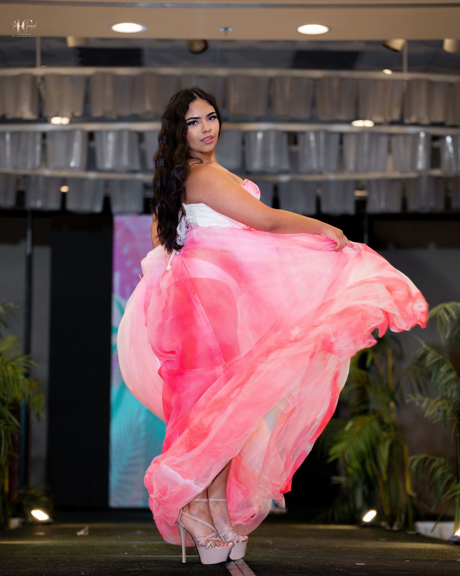 A model in a flowing coral pink dress poses dramatically against an industrial backdrop.