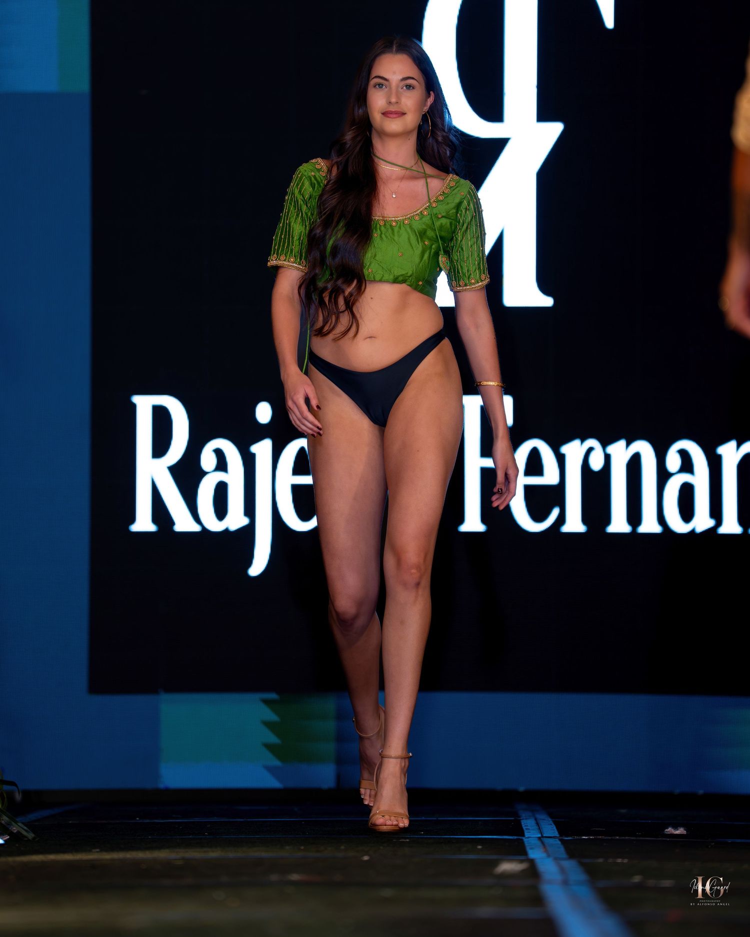 Model walks the runway in a black swimsuit during a fashion show with dark backdrop and dramatic lighting.