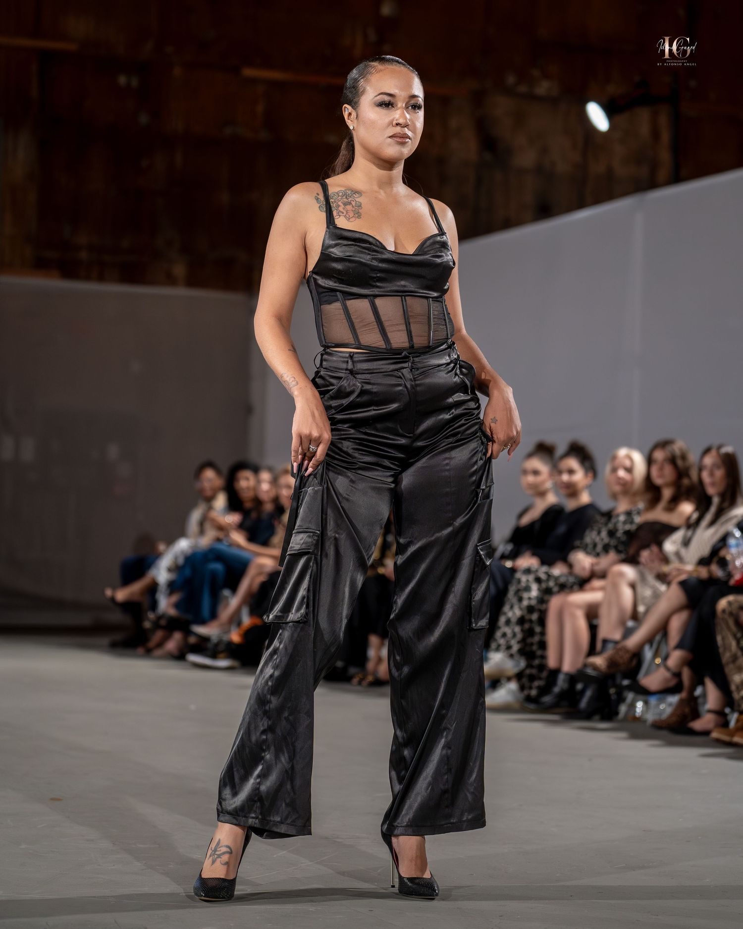 Model walks runway in flowing black evening gown during fashion show with audience in background.