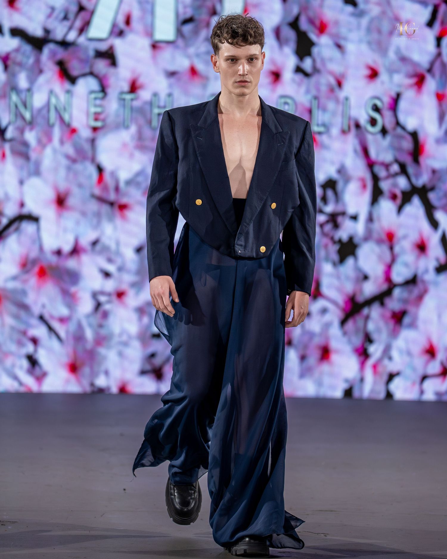 Fashion model walks runway in navy double-breasted blazer and wide-leg trousers against floral backdrop.