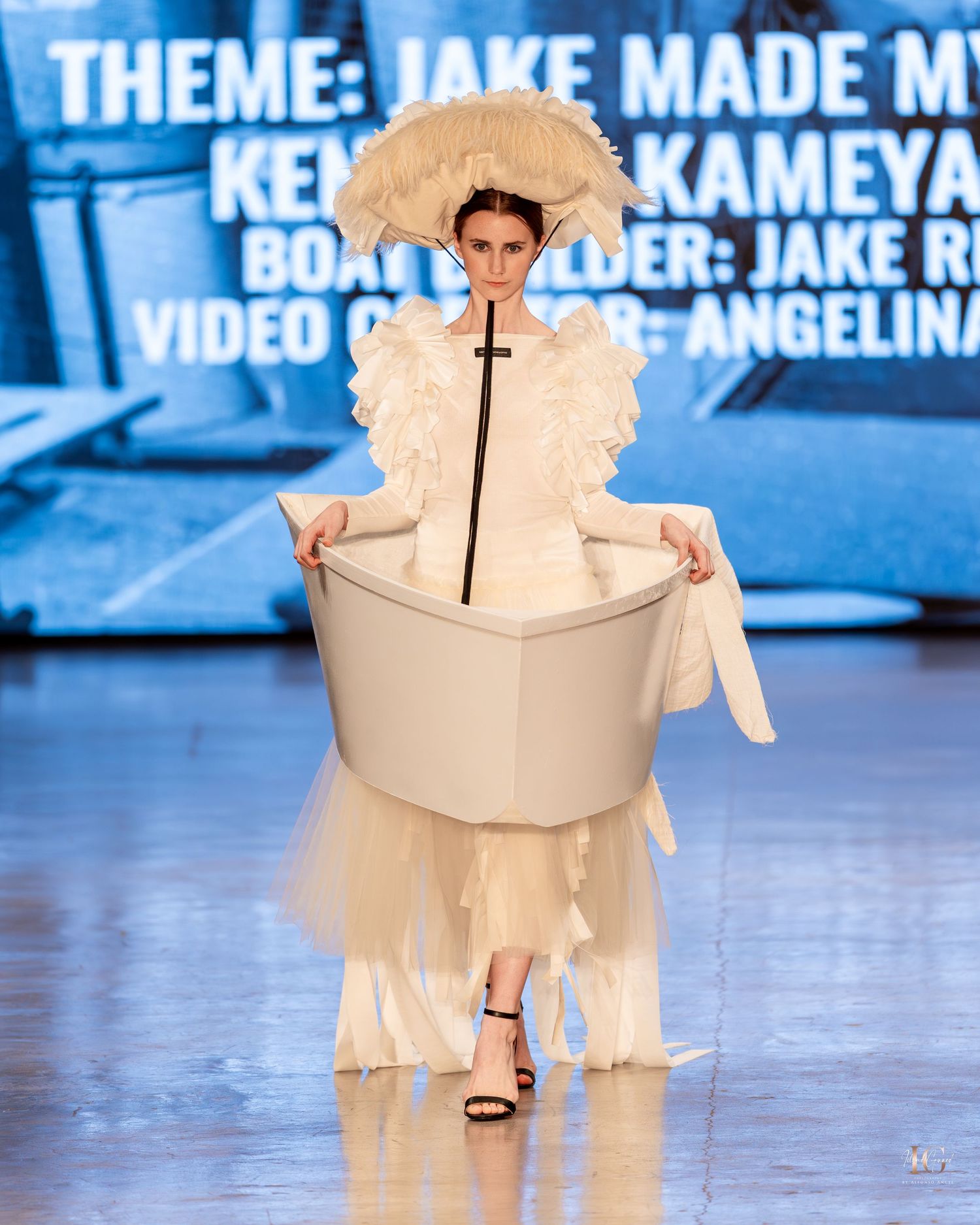 A fashion model wearing an elaborate cream-colored dress designed to look like a teacup on a runway with blue backdrop.
