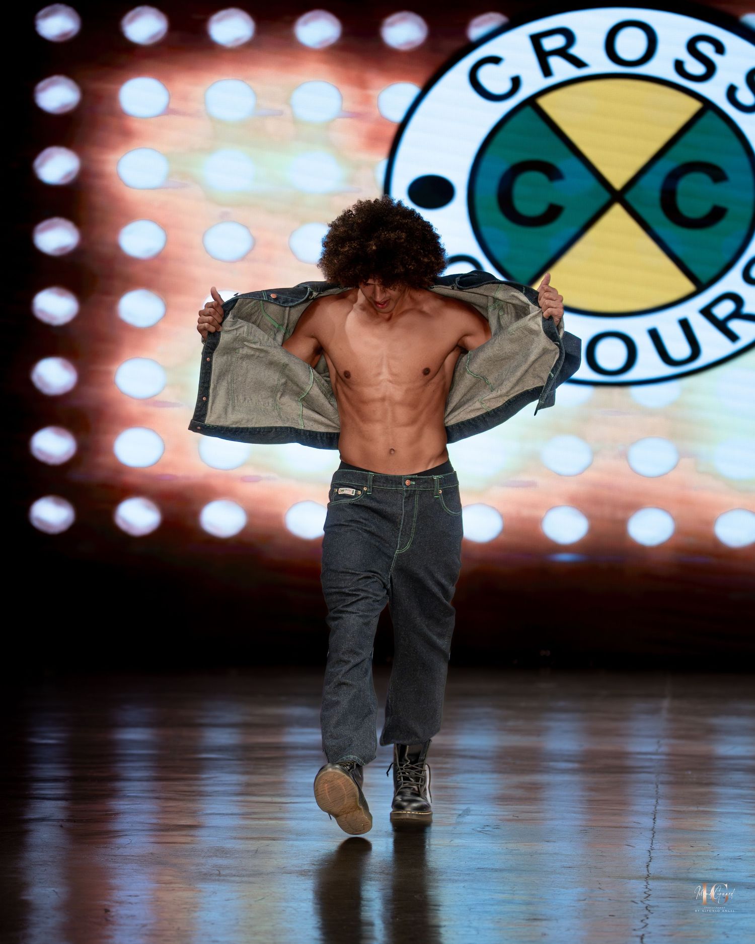 A performer in jeans and a beige jacket dances across a stage with illuminated circular lights in the background.