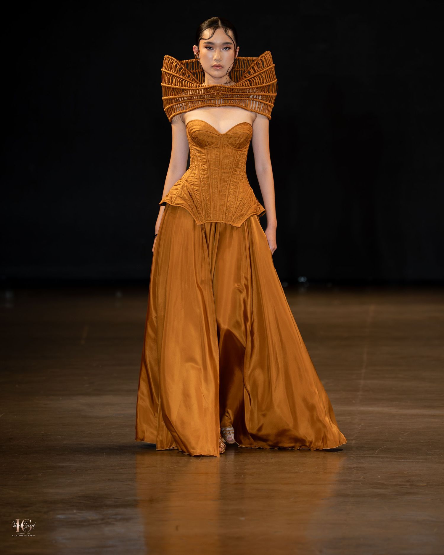 A model walks the runway wearing a flowing burnt orange gown with dramatic ruffled shoulders at a fashion show.