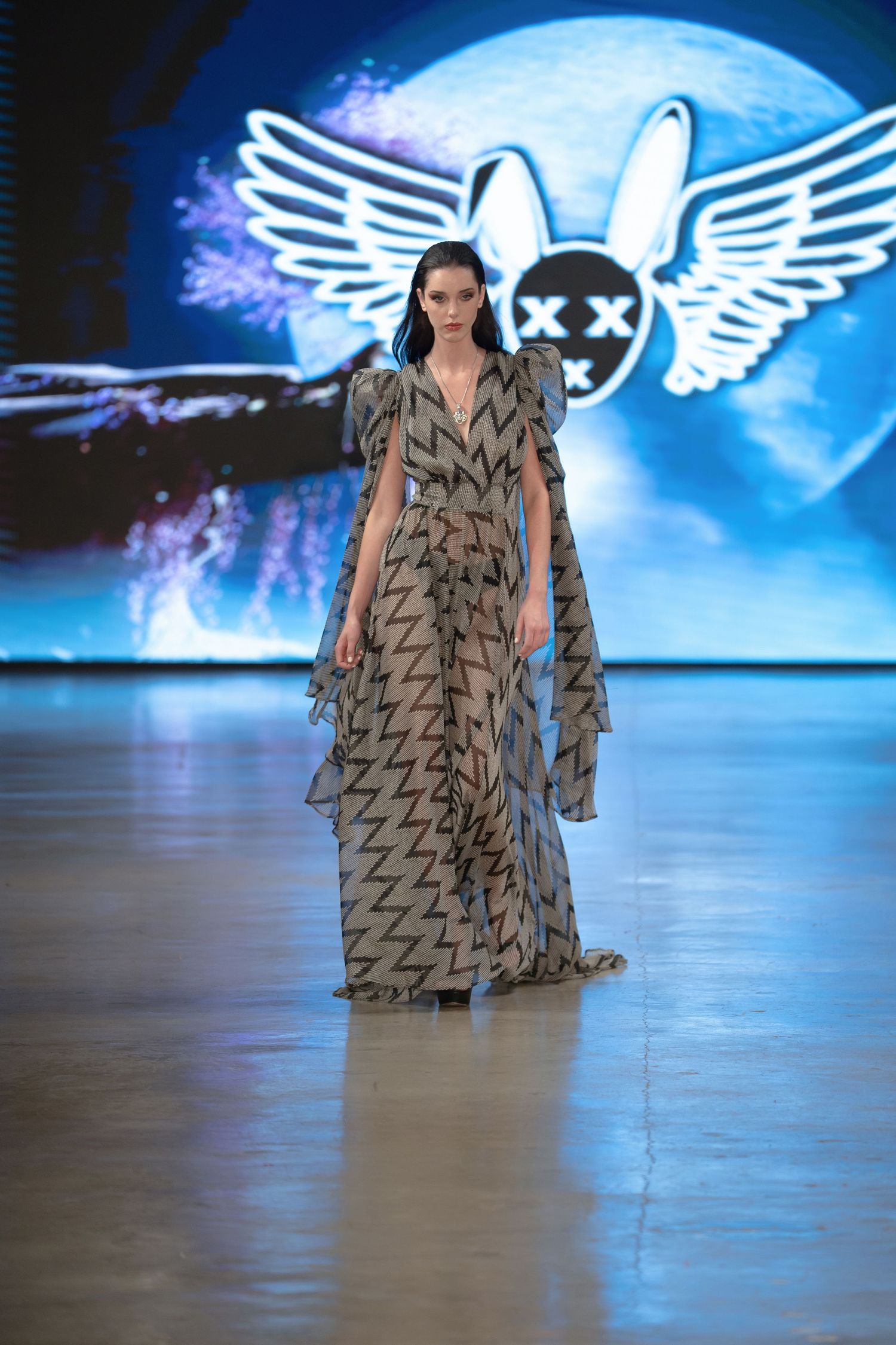 Model walks runway in flowing taupe dress with geometric print pattern at fashion show with illuminated winged logo backdrop.