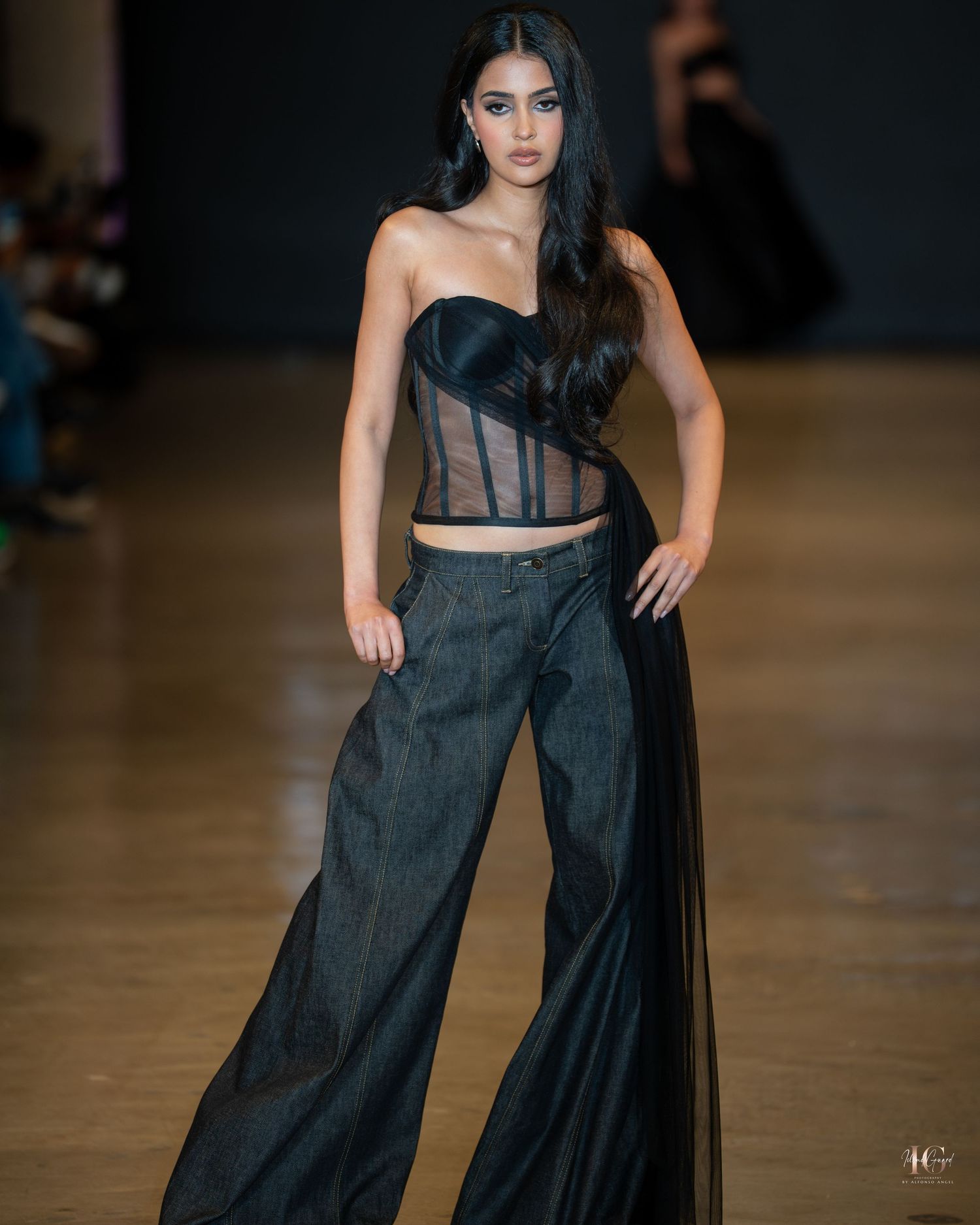 Fashion model walks runway in sheer black evening gown with corset detail at luxury fashion show.