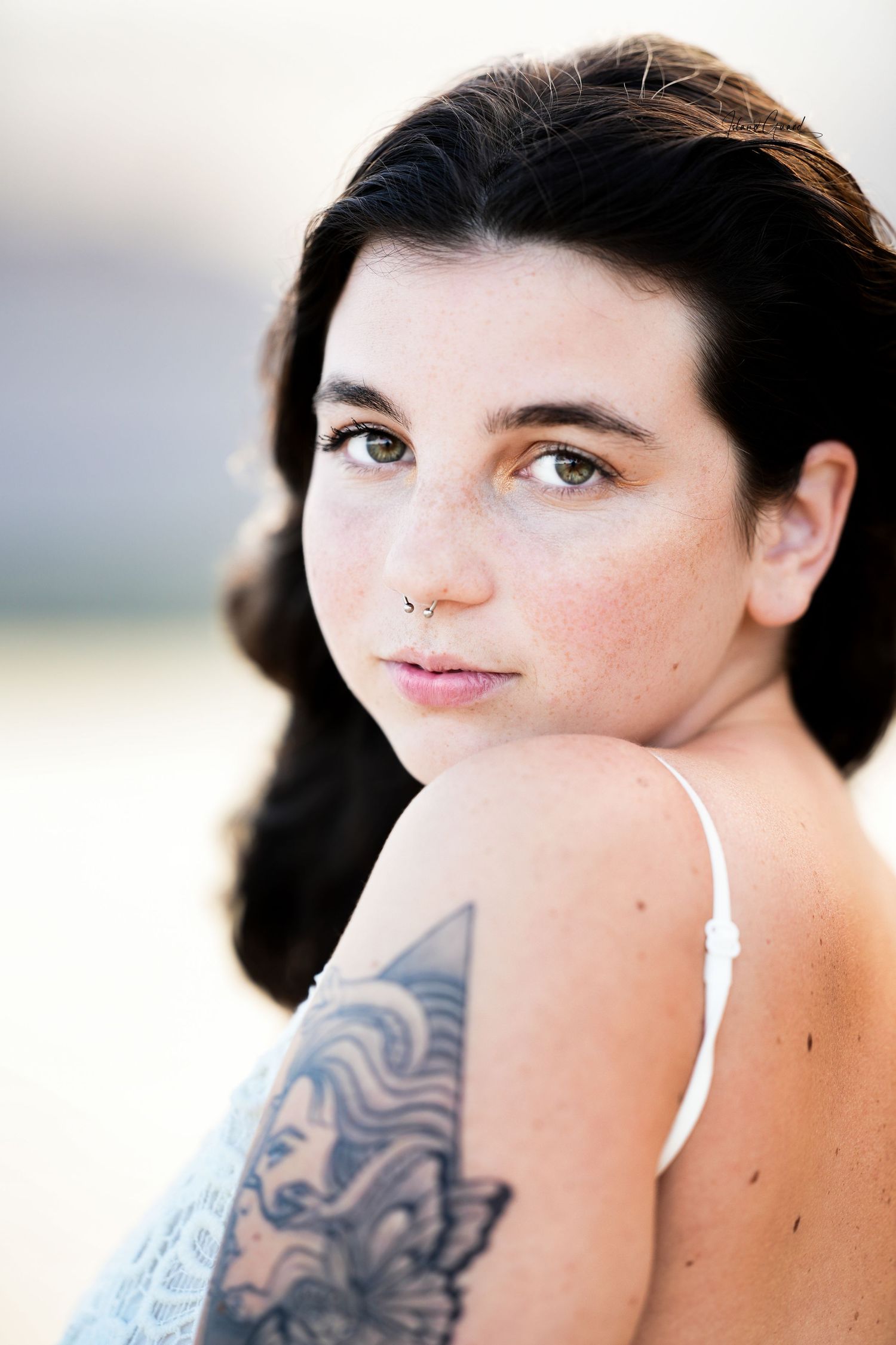 Close-up portrait of someone with dark hair wearing a white strap top and displaying an intricate floral arm tattoo.