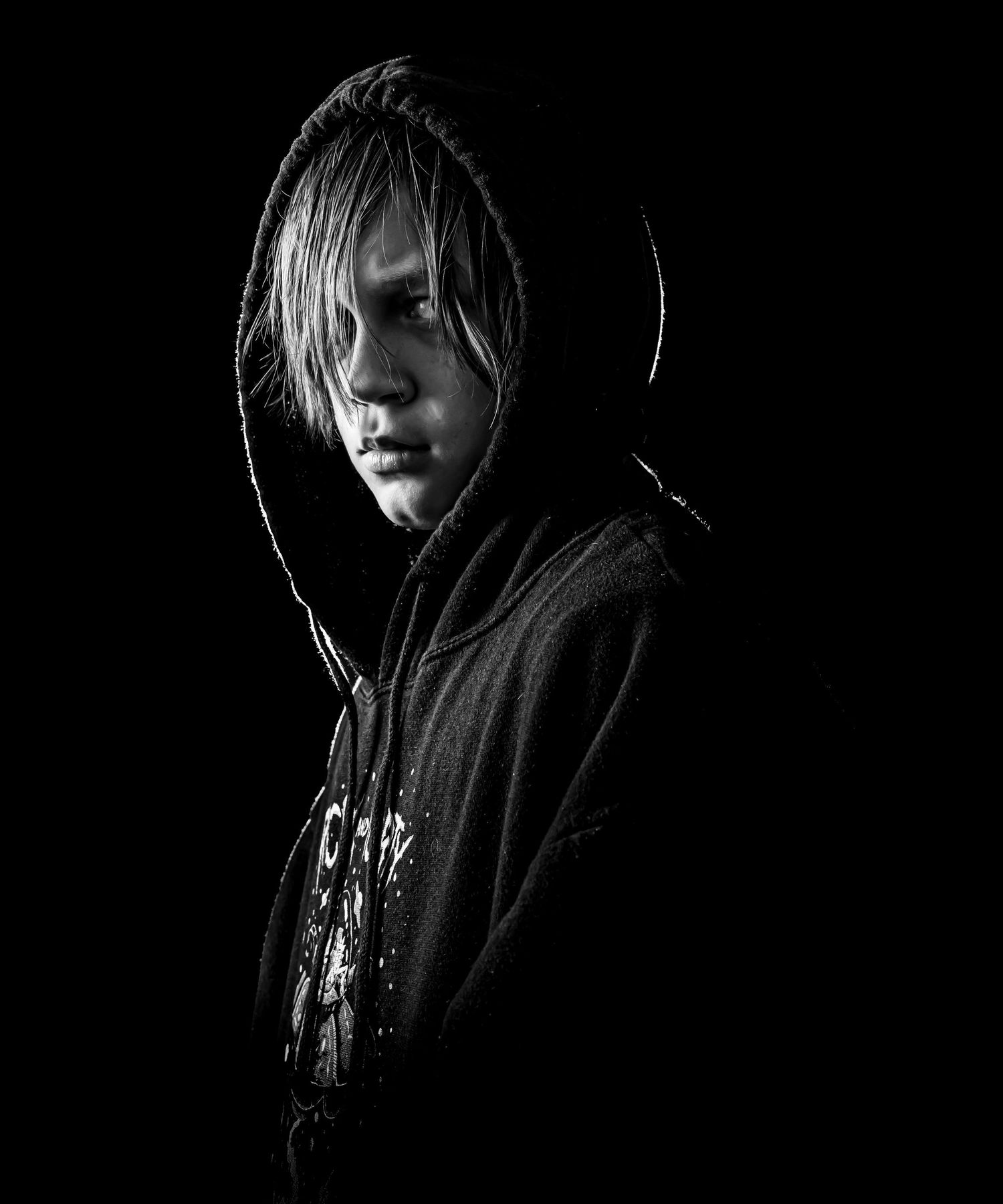 Dramatic black and white portrait showing figure in dark hoodie with hair falling across face against dark background.