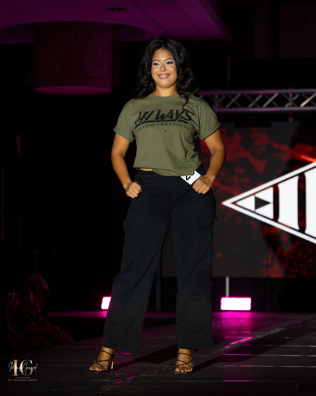 Model showcases olive green top and black pants on dark runway with pink accent lights.