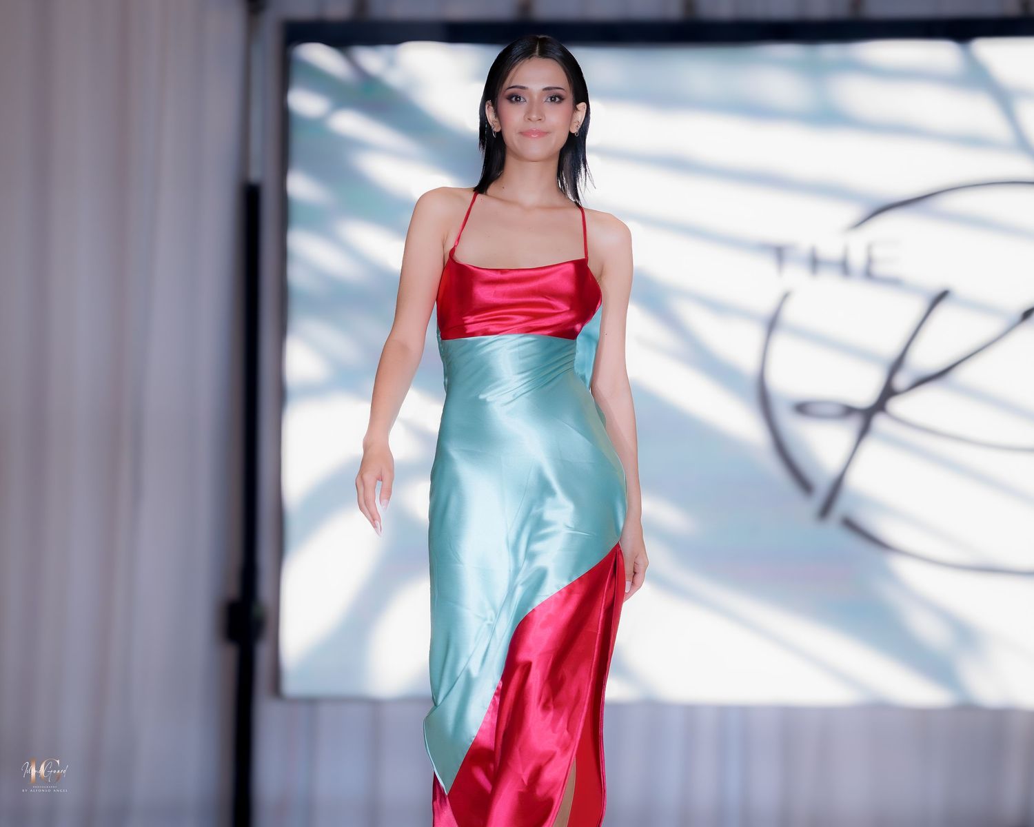 Model showcases red and turquoise evening gown on catwalk with dramatic lighting.