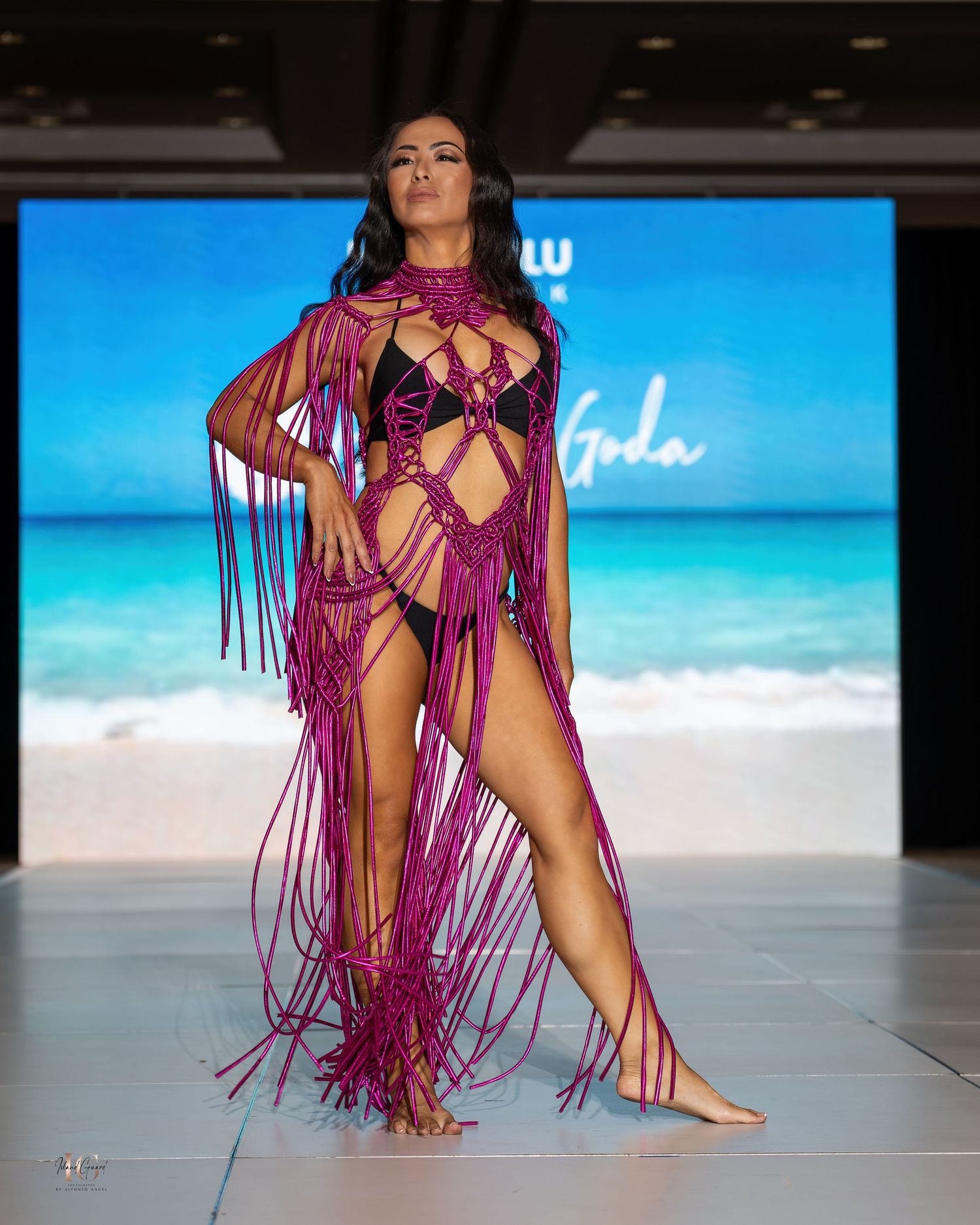 Magenta fringed swimwear with cutout details presented against beach backdrop at fashion show.