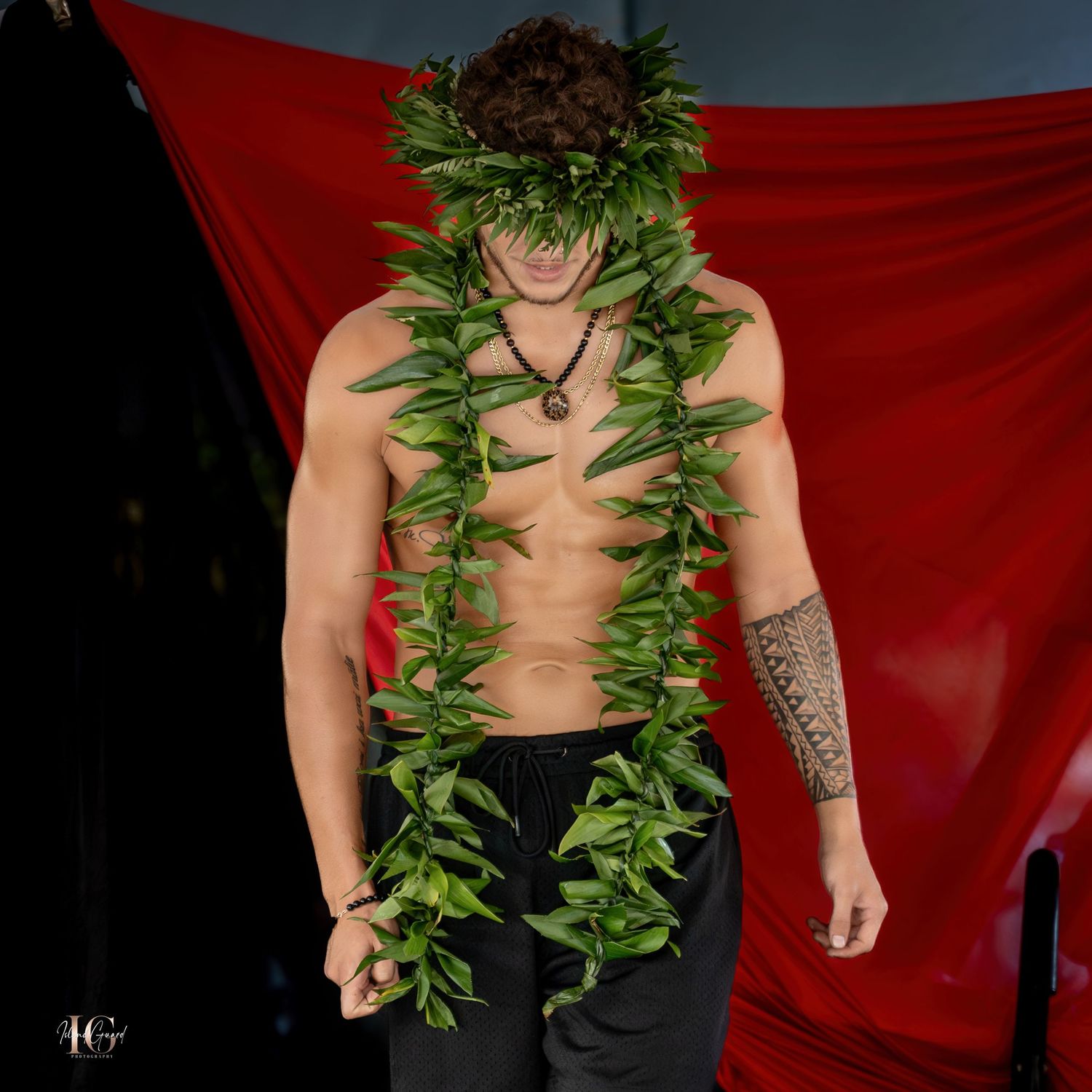 Performance artist wearing black pants and green leaf lei on stage with red backdrop.