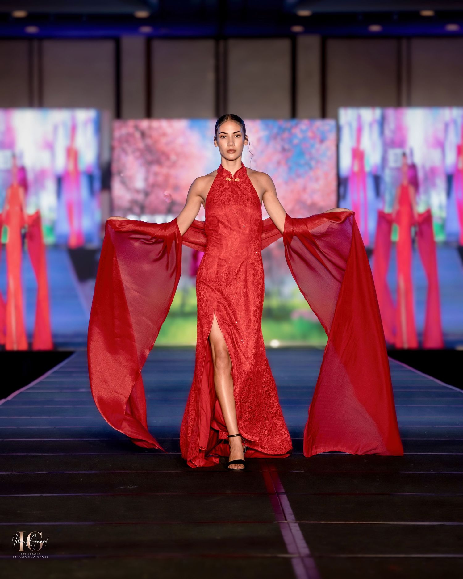 Model showcases flowing red evening gown with dramatic cape on illuminated fashion runway.