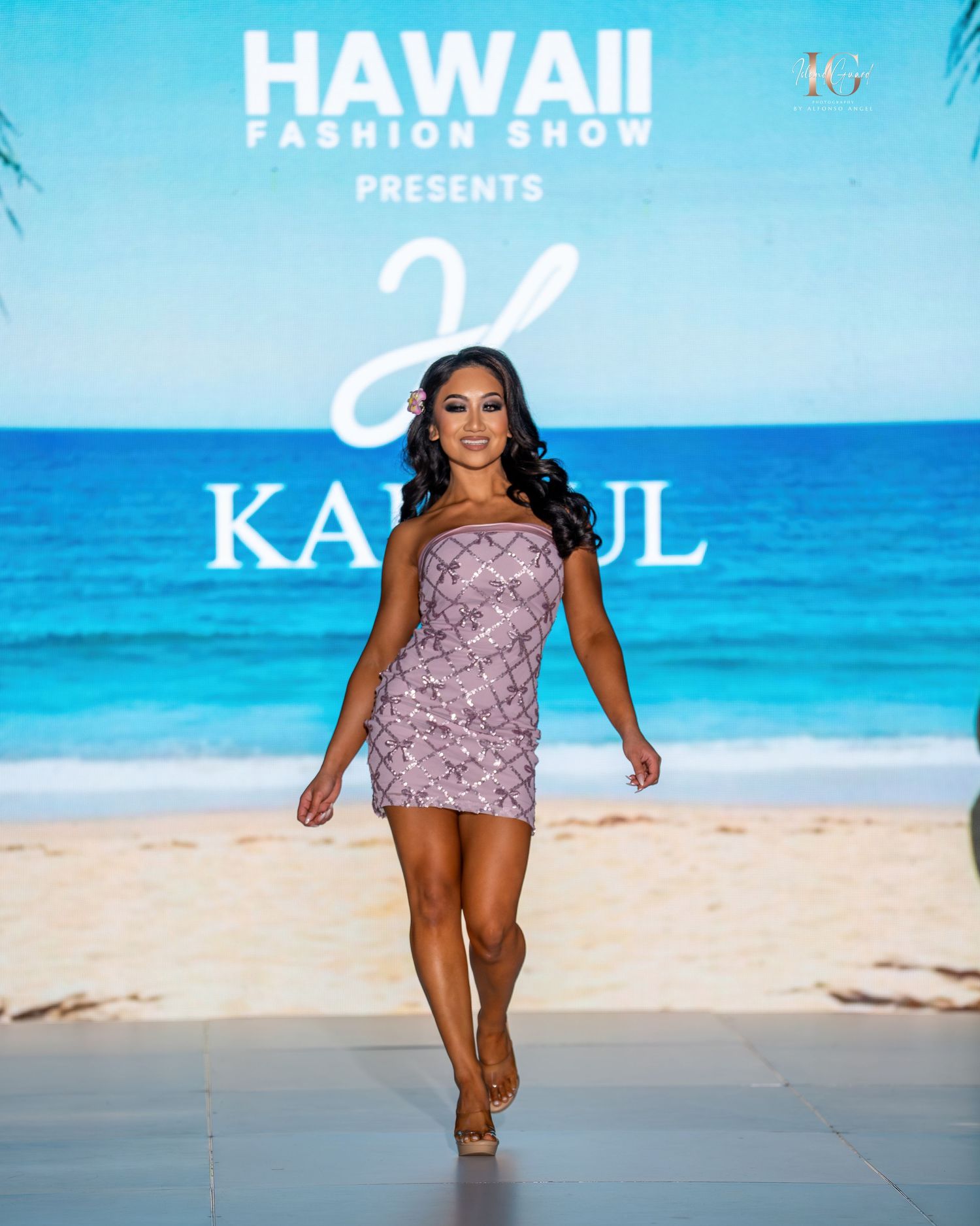 Model walks the runway in a lavender sequined dress at a Hawaii fashion show with ocean backdrop.