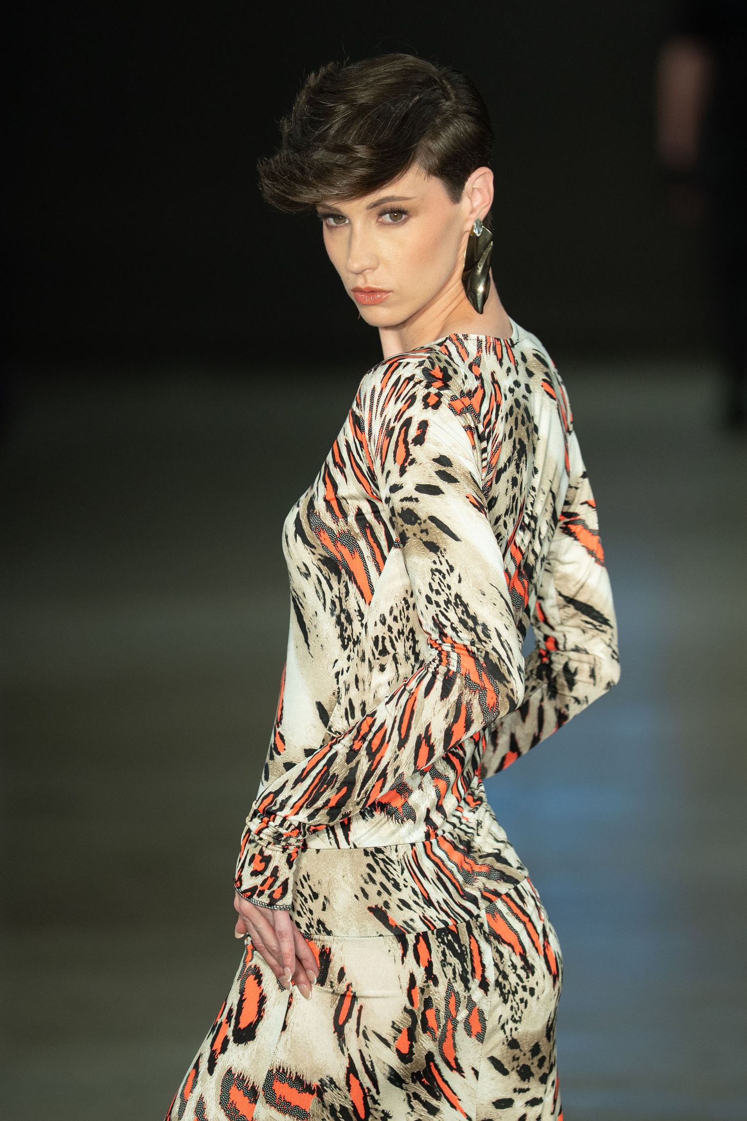 Model walks runway in flowing abstract print dress with orange and black pattern against dark background at fashion show.