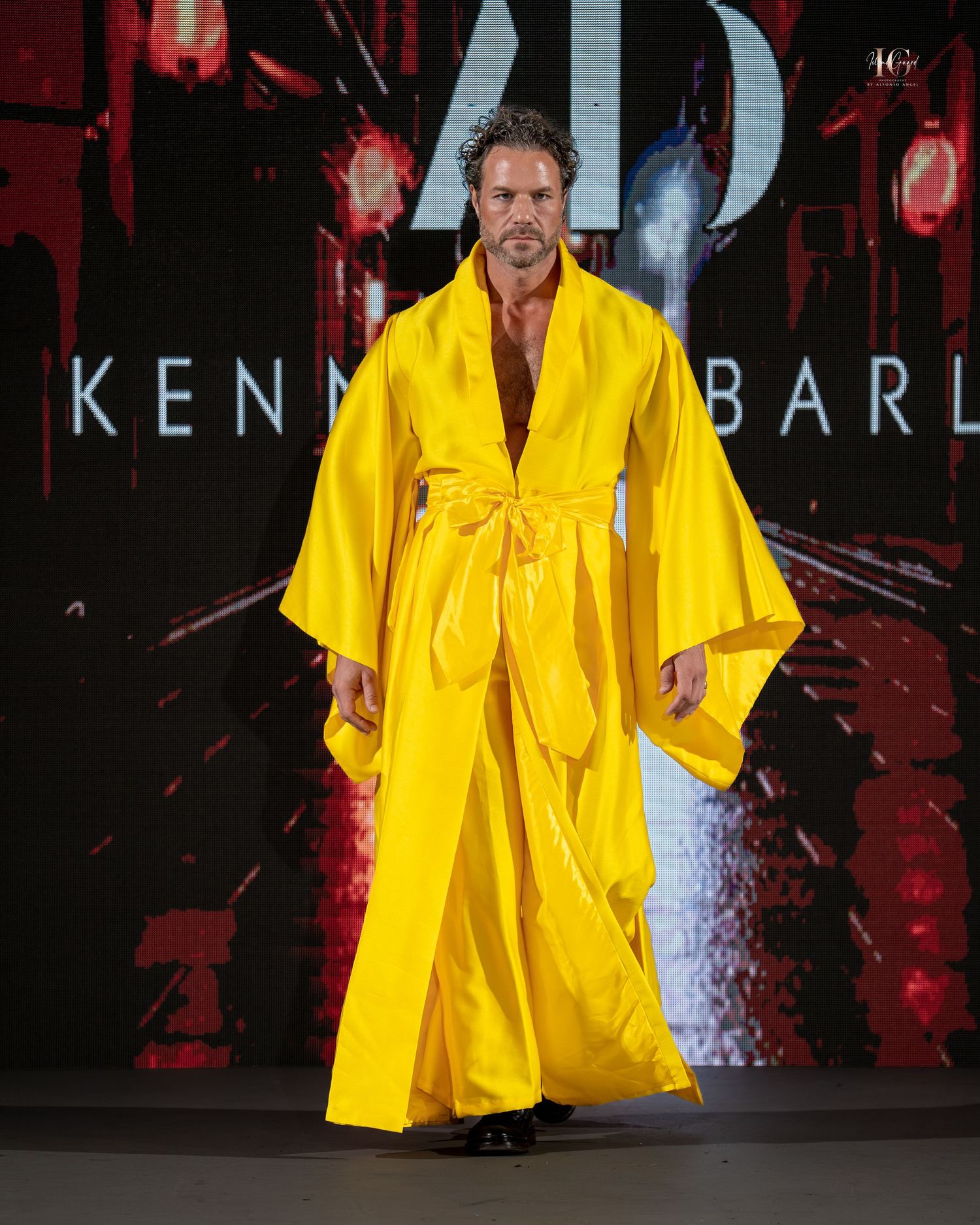 Performer in bright yellow outfit stands on stage with dramatic red lighting and dark background.