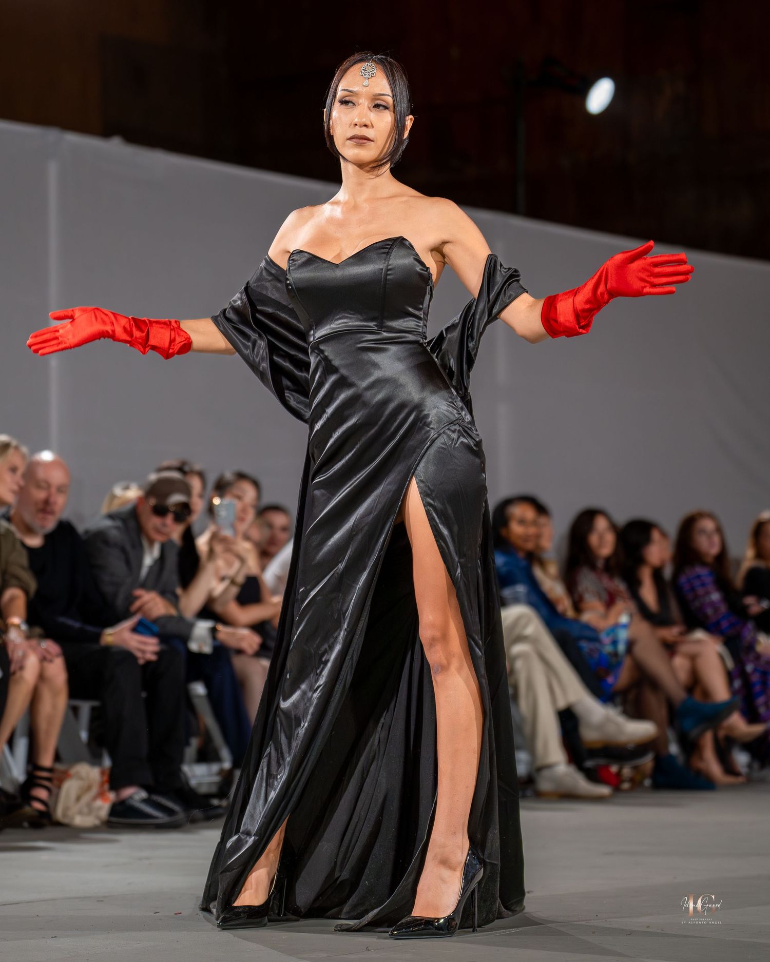Model walks runway in elegant black satin gown with dramatic red gloves during fashion show.
