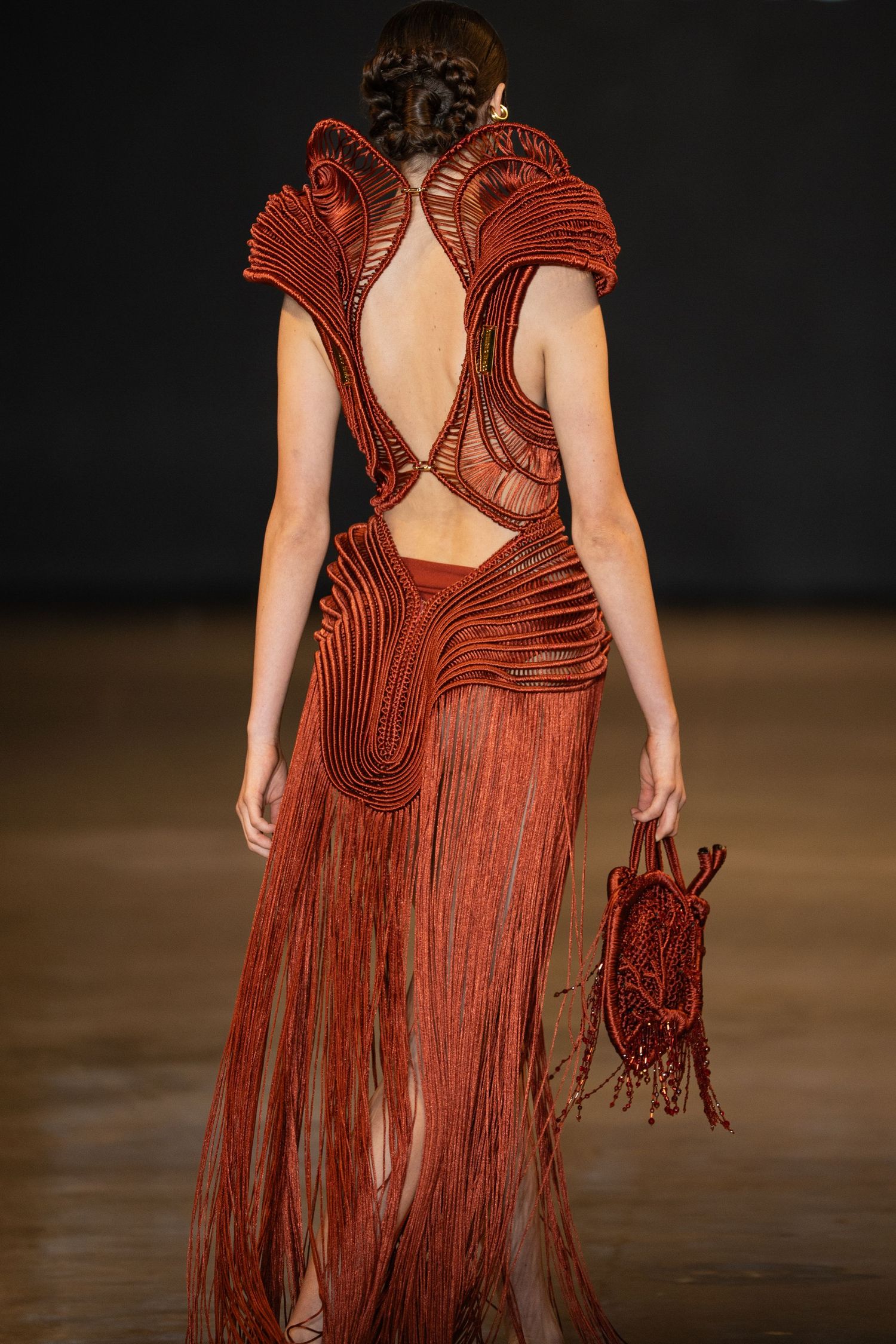Runway model wearing an avant-garde rust-colored dress with intricate pleating and dramatic cutout back details.