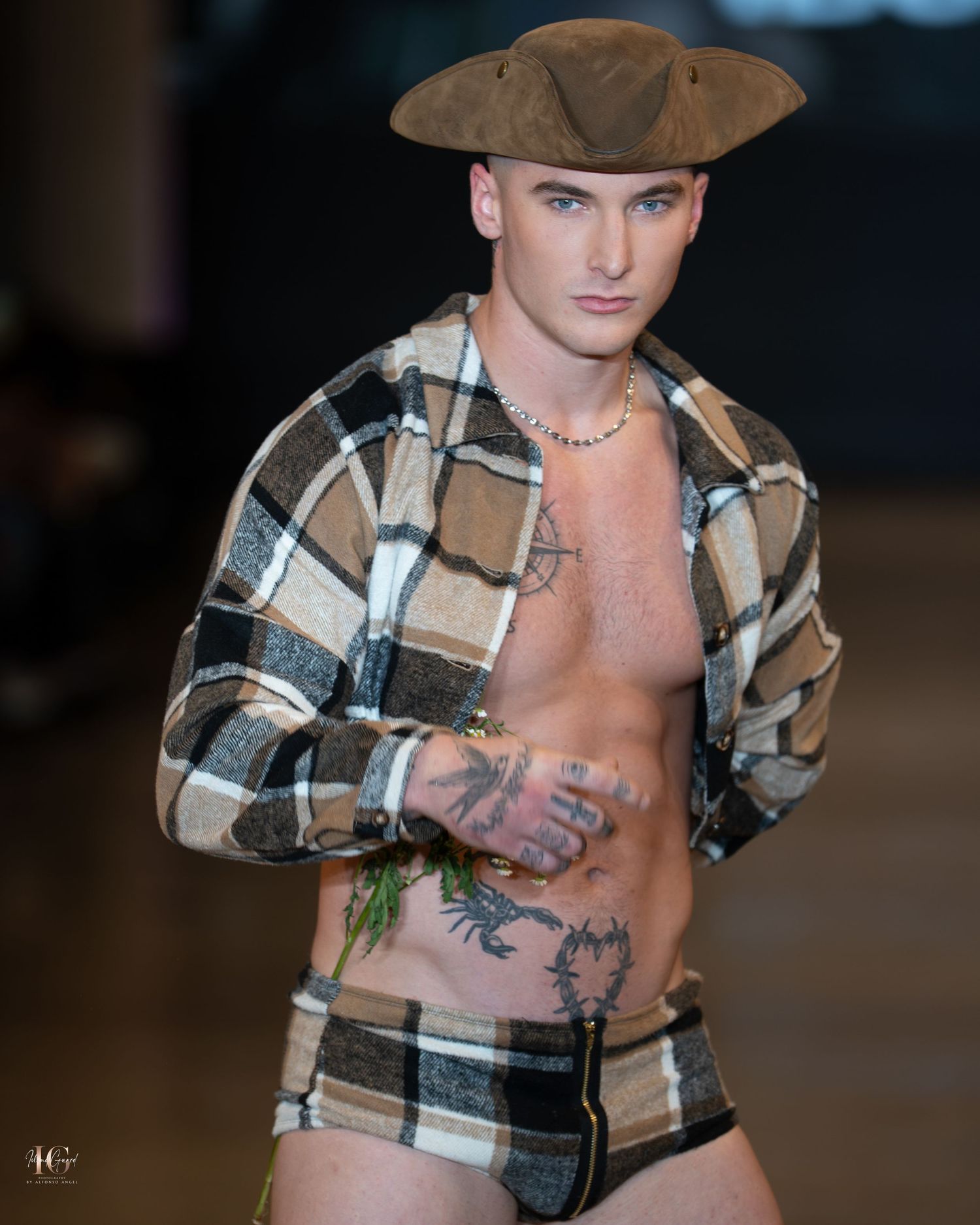 Model walks runway in brown plaid flannel shirt with open chest showing tattoos during fashion show presentation.