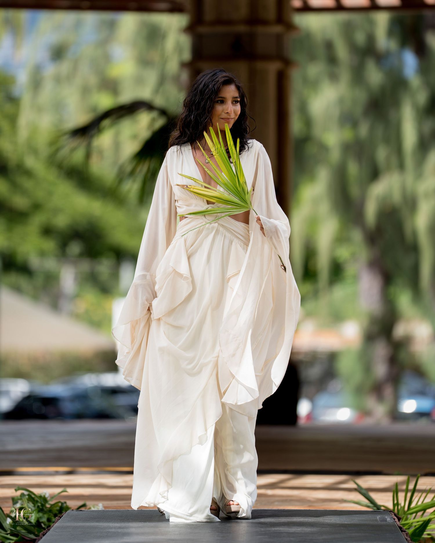 A person in a flowing white dress stands elegantly in a garden setting with dappled sunlight filtering through trees.