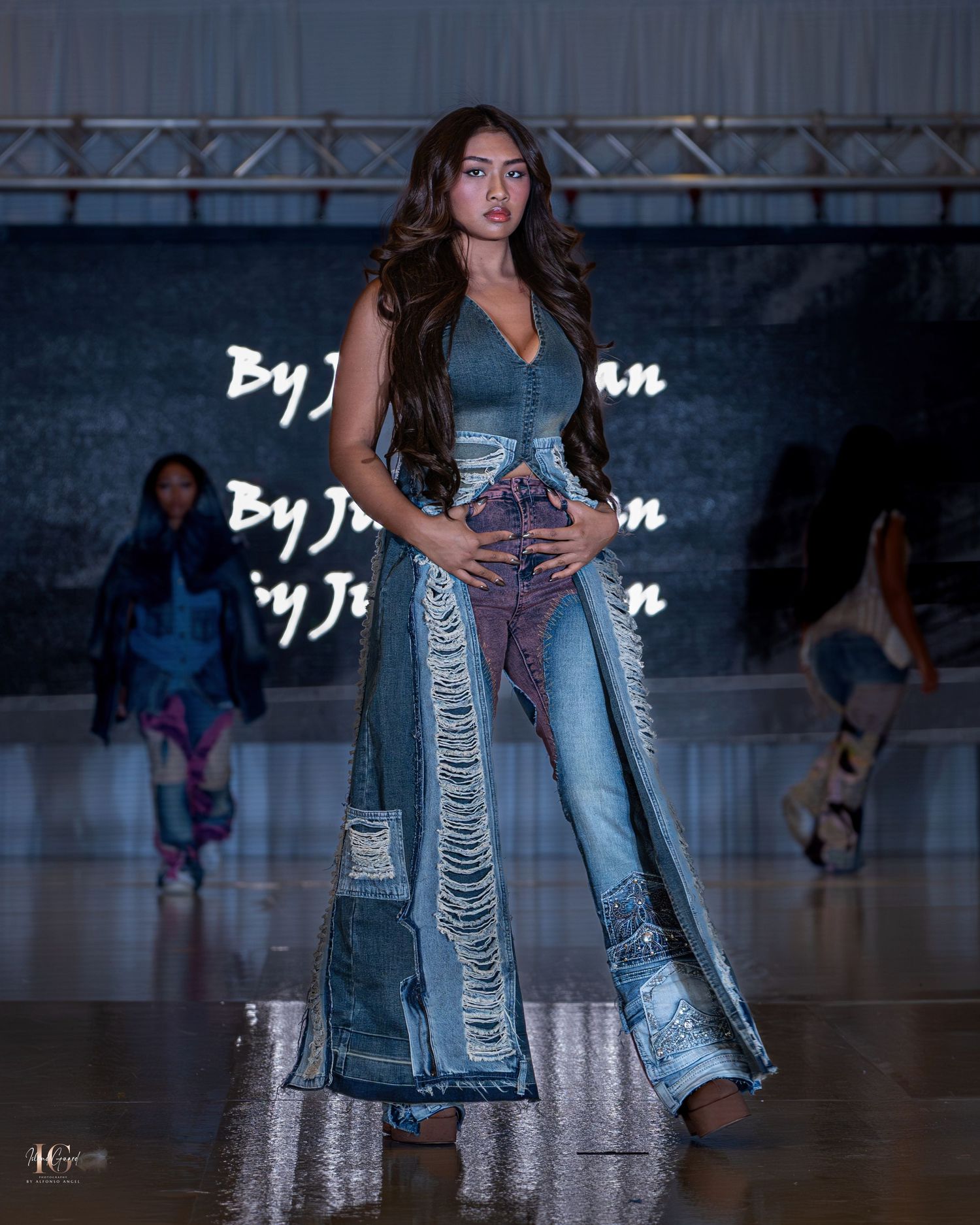 Model walks runway wearing distressed denim jeans and long denim vest at fashion show on reflective floor.