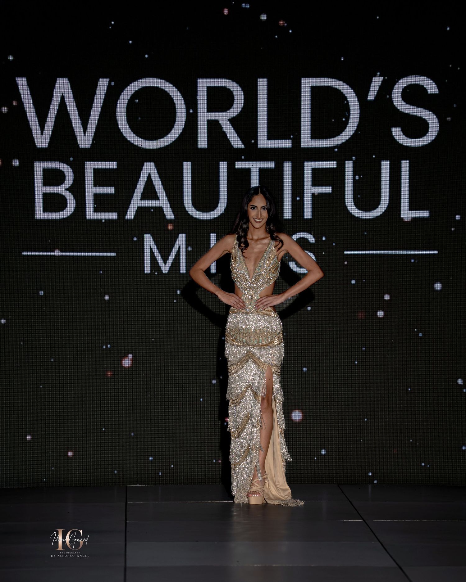 Model walks runway in elegant beige evening gown with plunging neckline at fashion show with illuminated backdrop.