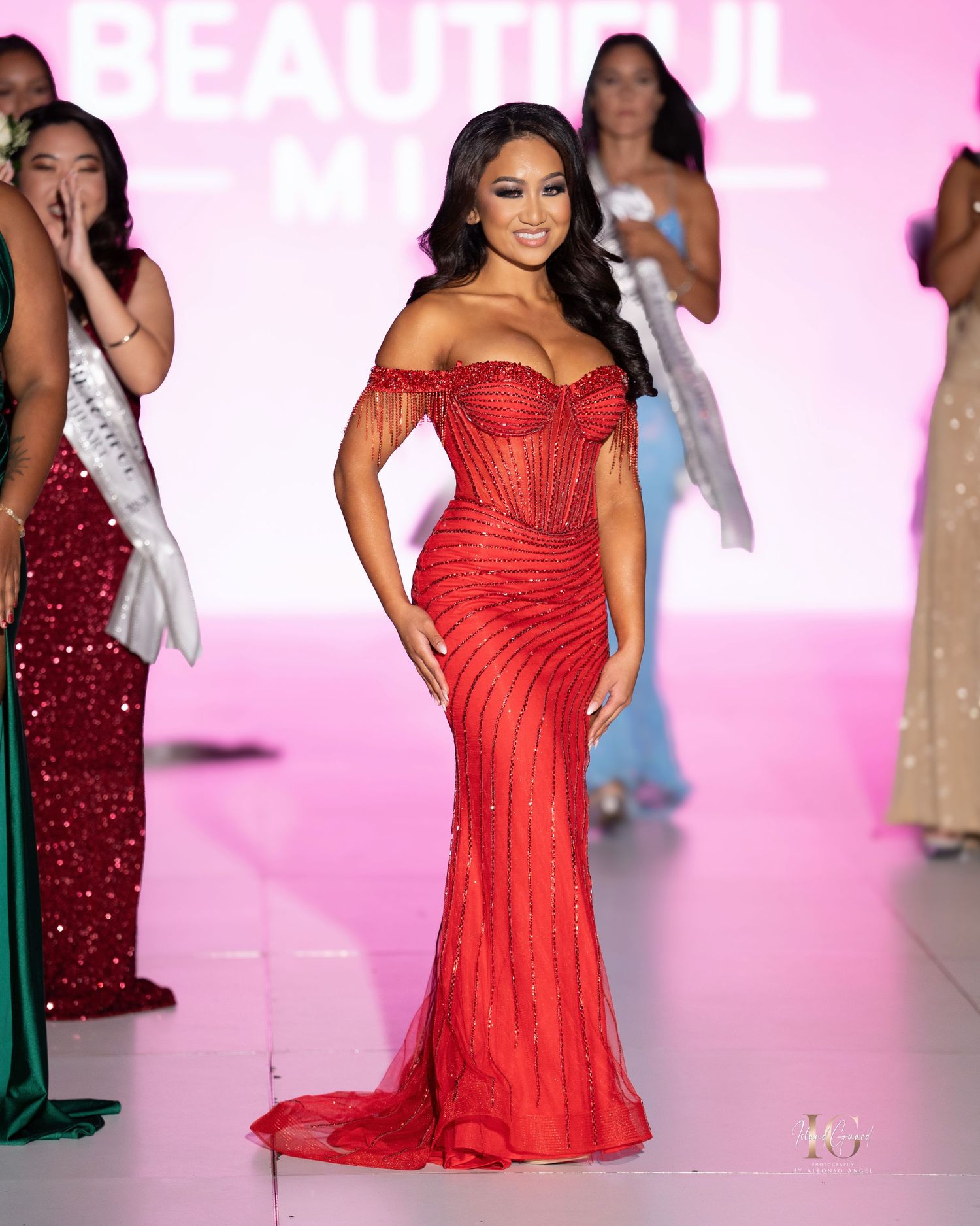Models walk the runway in red evening gowns during a glamorous fashion show with pink lighting.