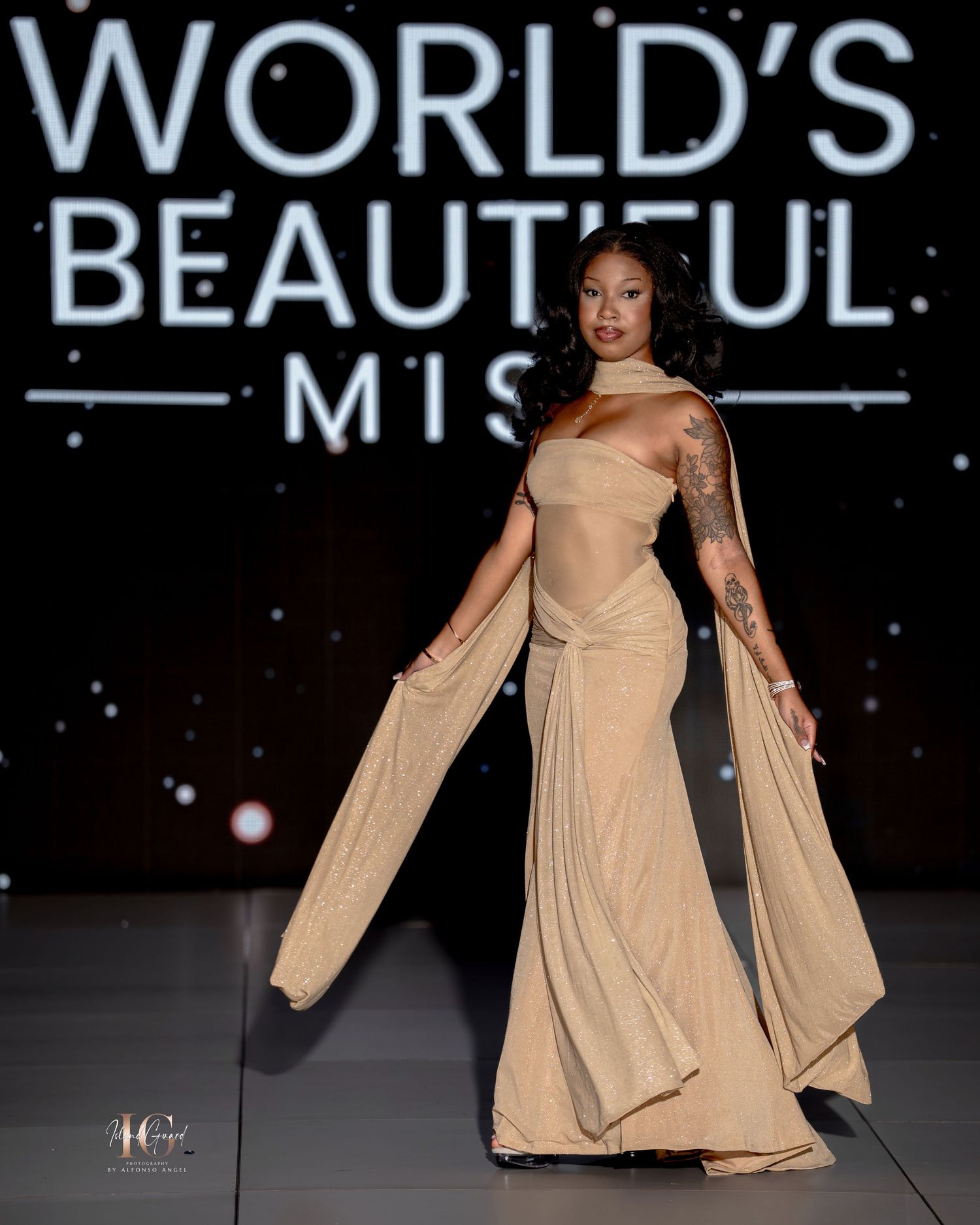 A model walks the runway in a flowing beige gown with dramatic cape sleeves at the World's Beautiful pageant show.