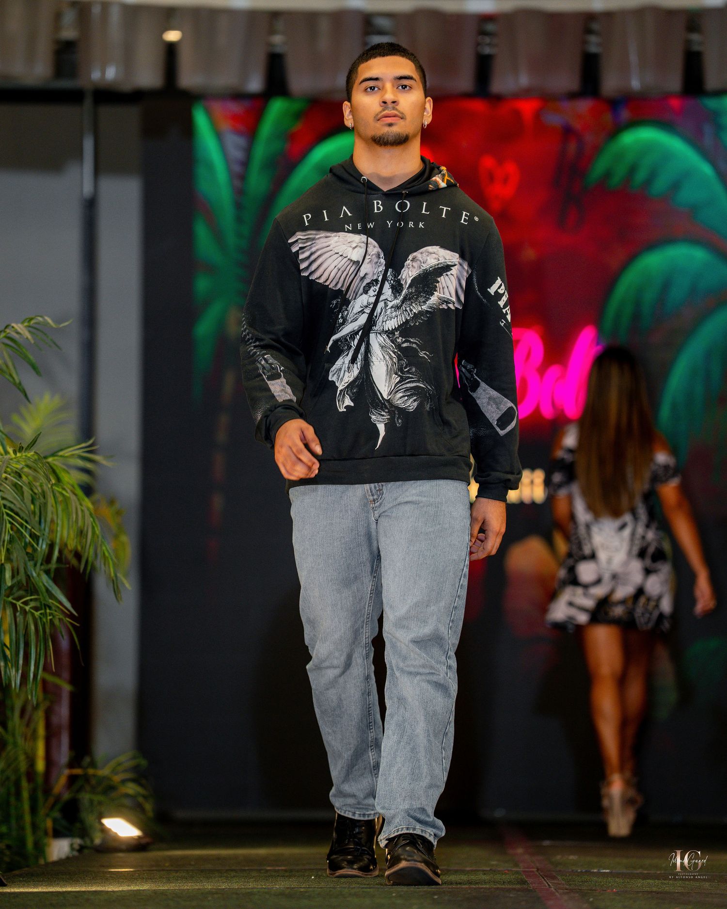 Model walks down runway wearing black graphic t-shirt and light gray jeans during fashion show.
