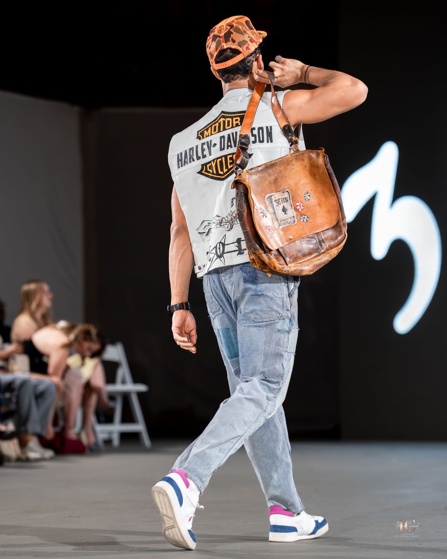 Model walks runway wearing Harley-Davidson t-shirt, distressed jeans and sneakers while carrying brown leather bag.