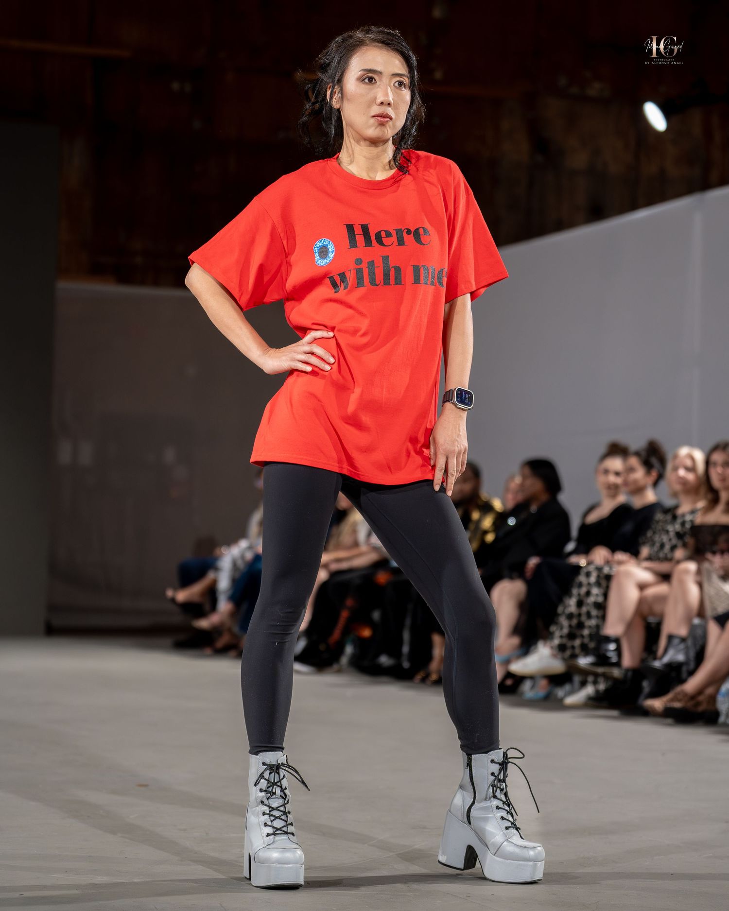 Model walks runway wearing red graphic t-shirt with leggings and platform boots at fashion show.