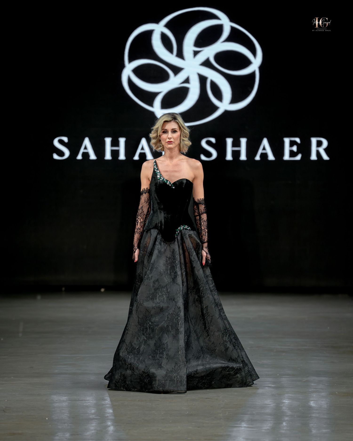 A model walks the runway in a dramatic black evening gown with a flowing cape at a fashion show.