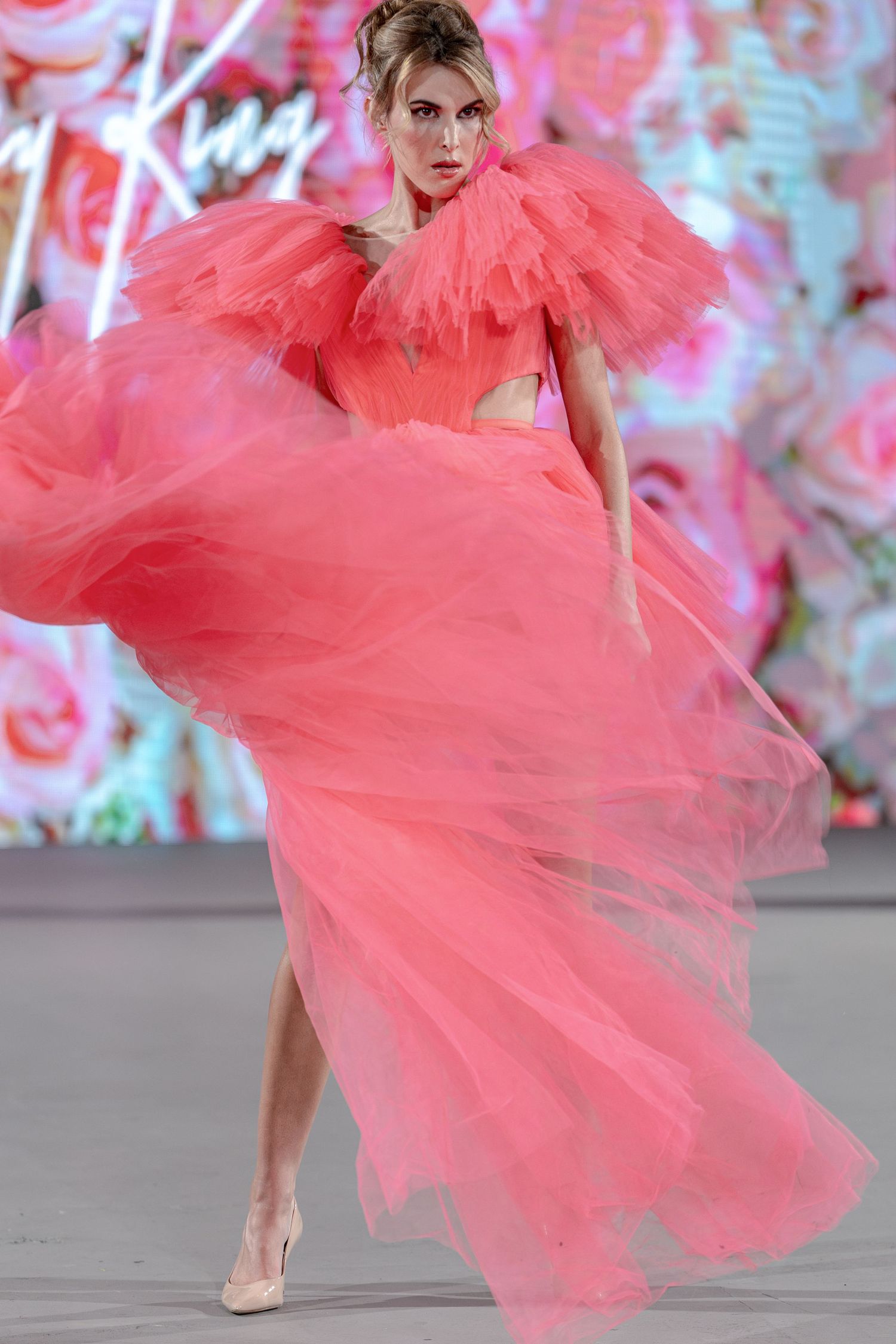 A model walks the runway in a voluminous coral tulle gown with dramatic ruffles against a pink floral backdrop.