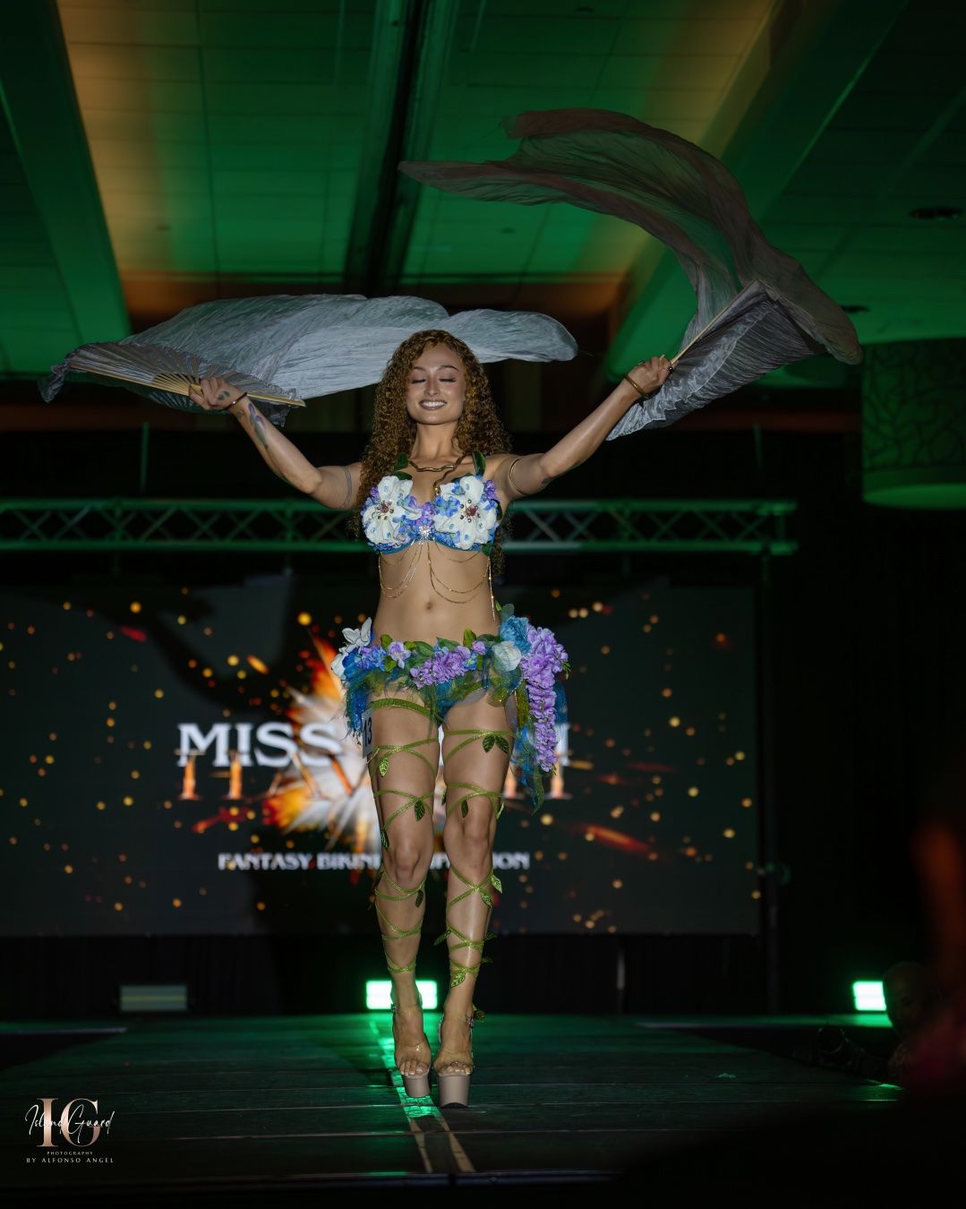 Fashion model walks the runway in shimmering silver lingerie with wing-style accessories under dramatic green lighting.