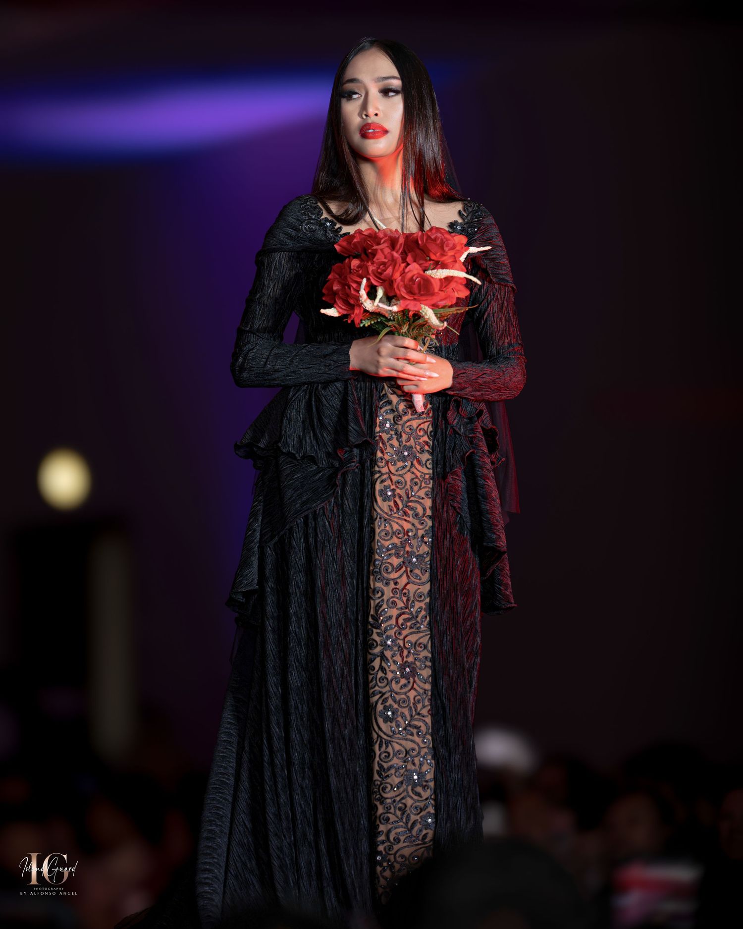 Fashion model in black lace evening gown carries vibrant red flowers on dark stage.