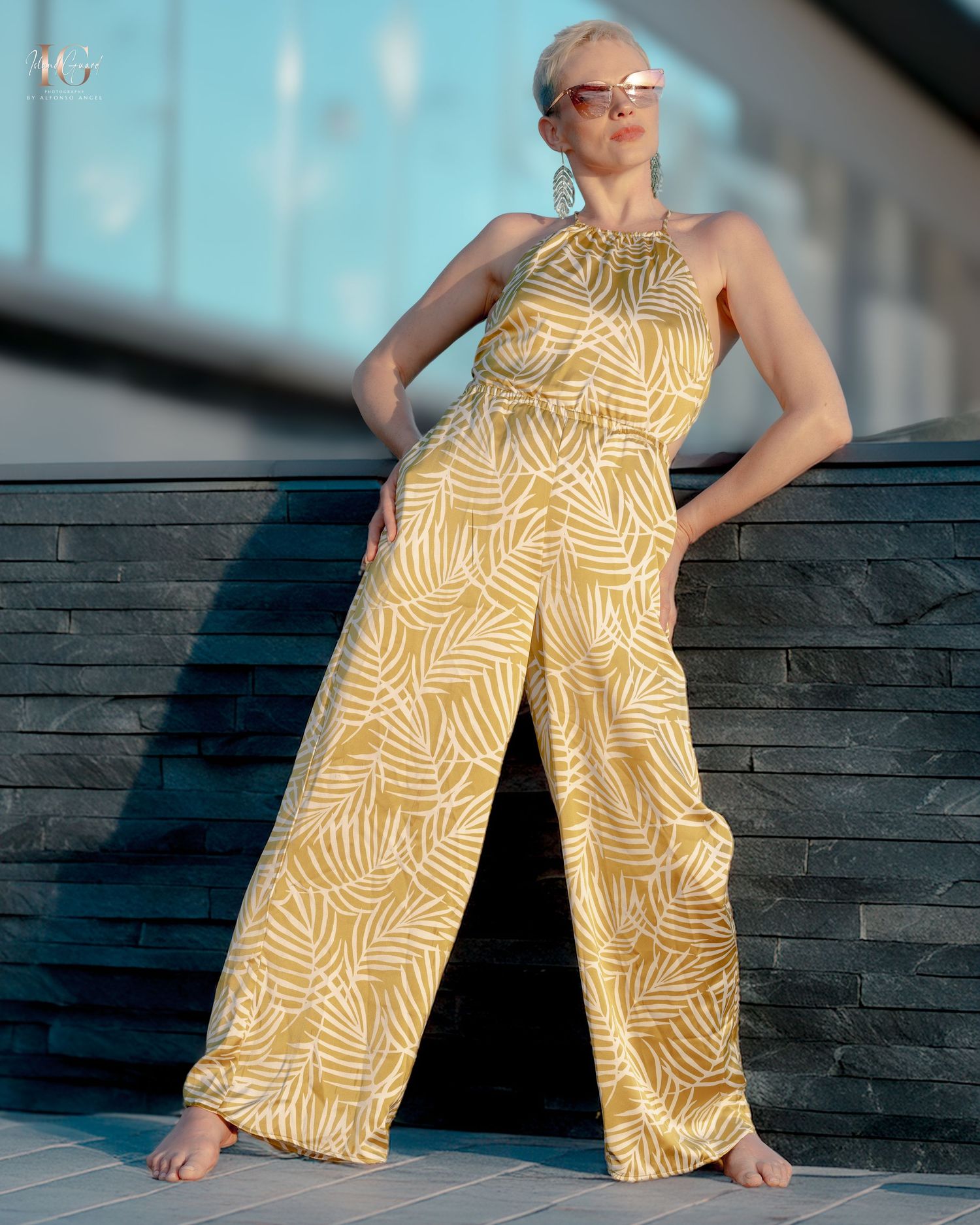 A model lounges on wooden decking wearing a yellow geometric print sundress in warm sunlight.