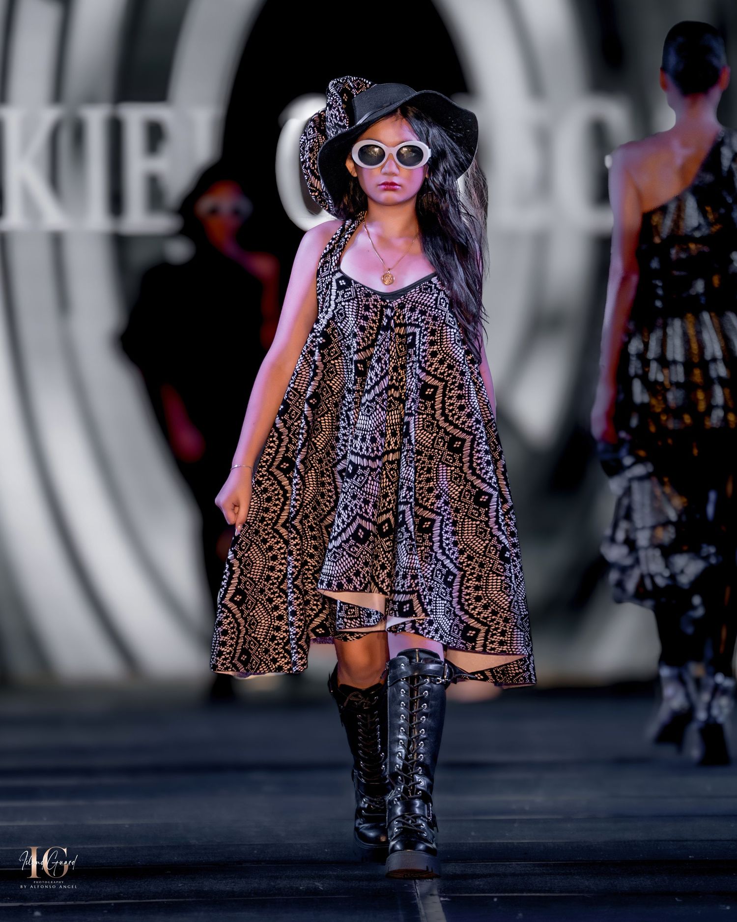 Runway model wearing loose-fitting printed dress and pink sunglasses with dramatic backdrop.
