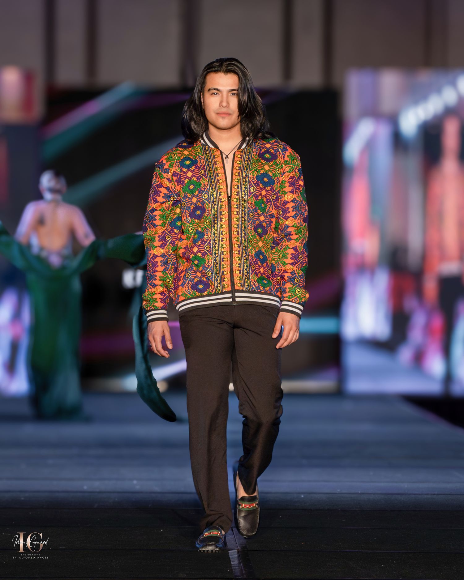 Embroidered ethnic jacket with colorful patterns worn over dark pants on fashion runway.