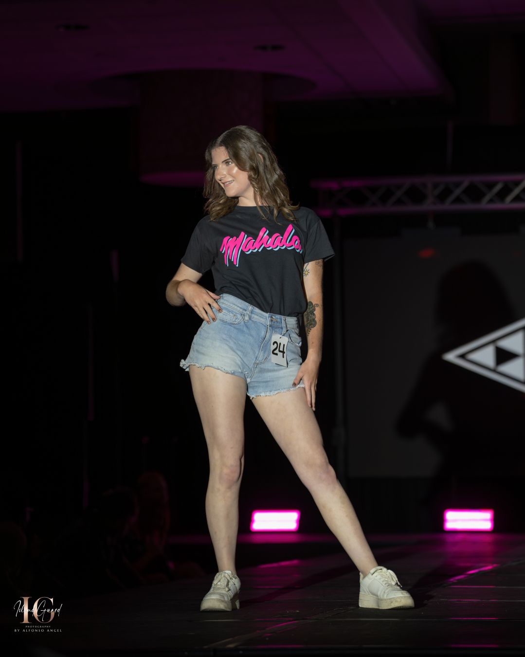 Model walks runway in black graphic t-shirt and denim shorts with pink stage lighting.