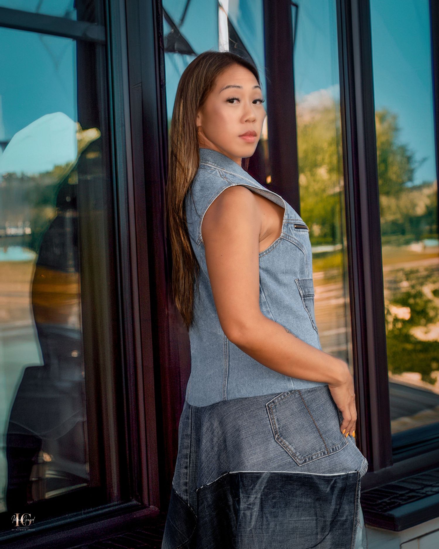 Person wearing a sleeveless denim top poses confidently against reflective windows with a turquoise backdrop.