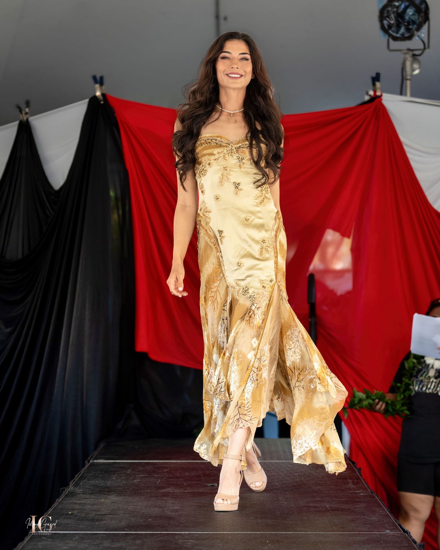 Model walks runway in shimmering gold evening gown against dramatic backdrop.