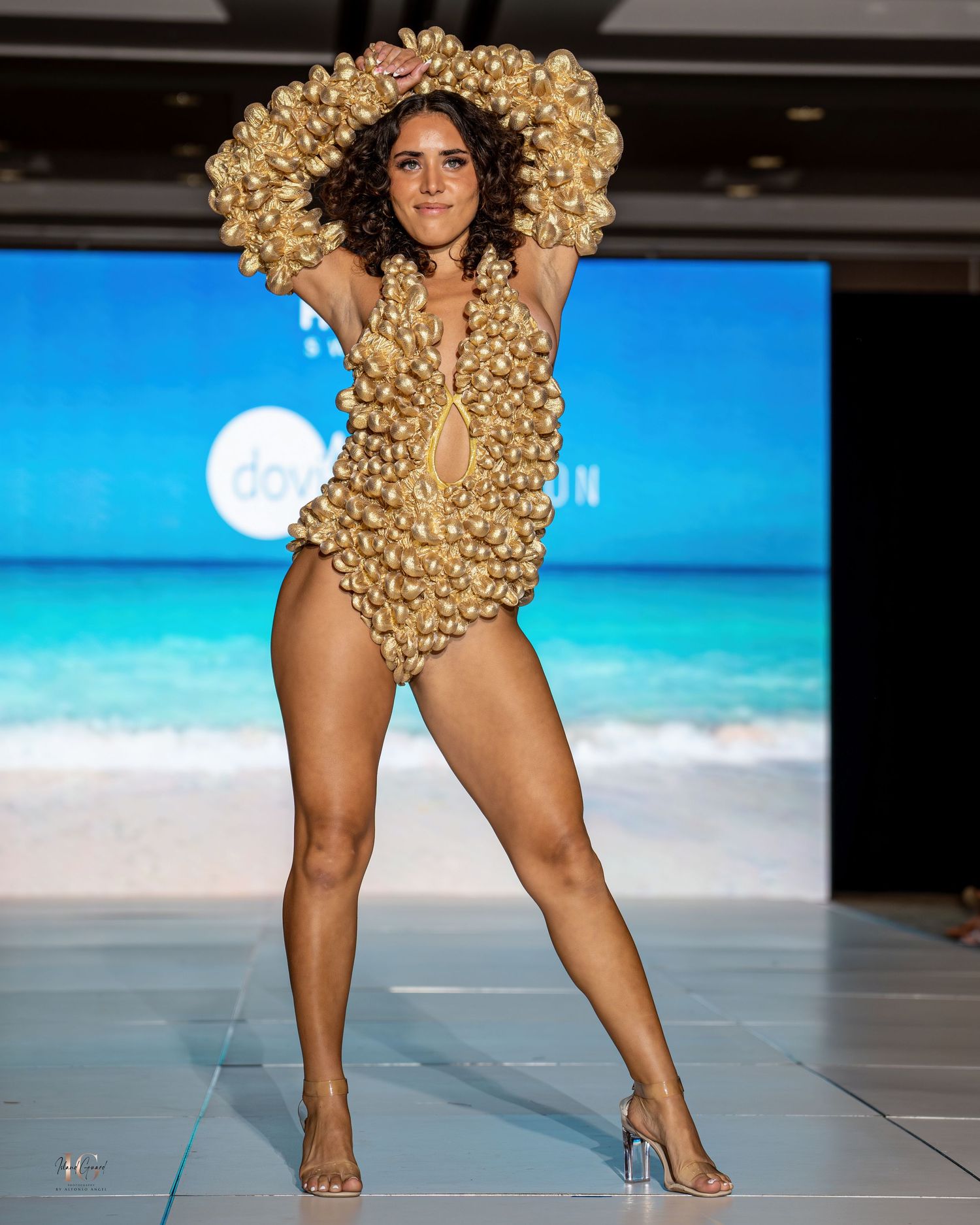 Model walks runway in avant-garde gold swimsuit with dramatic ruffled details at Honolulu Swim Week fashion show.
