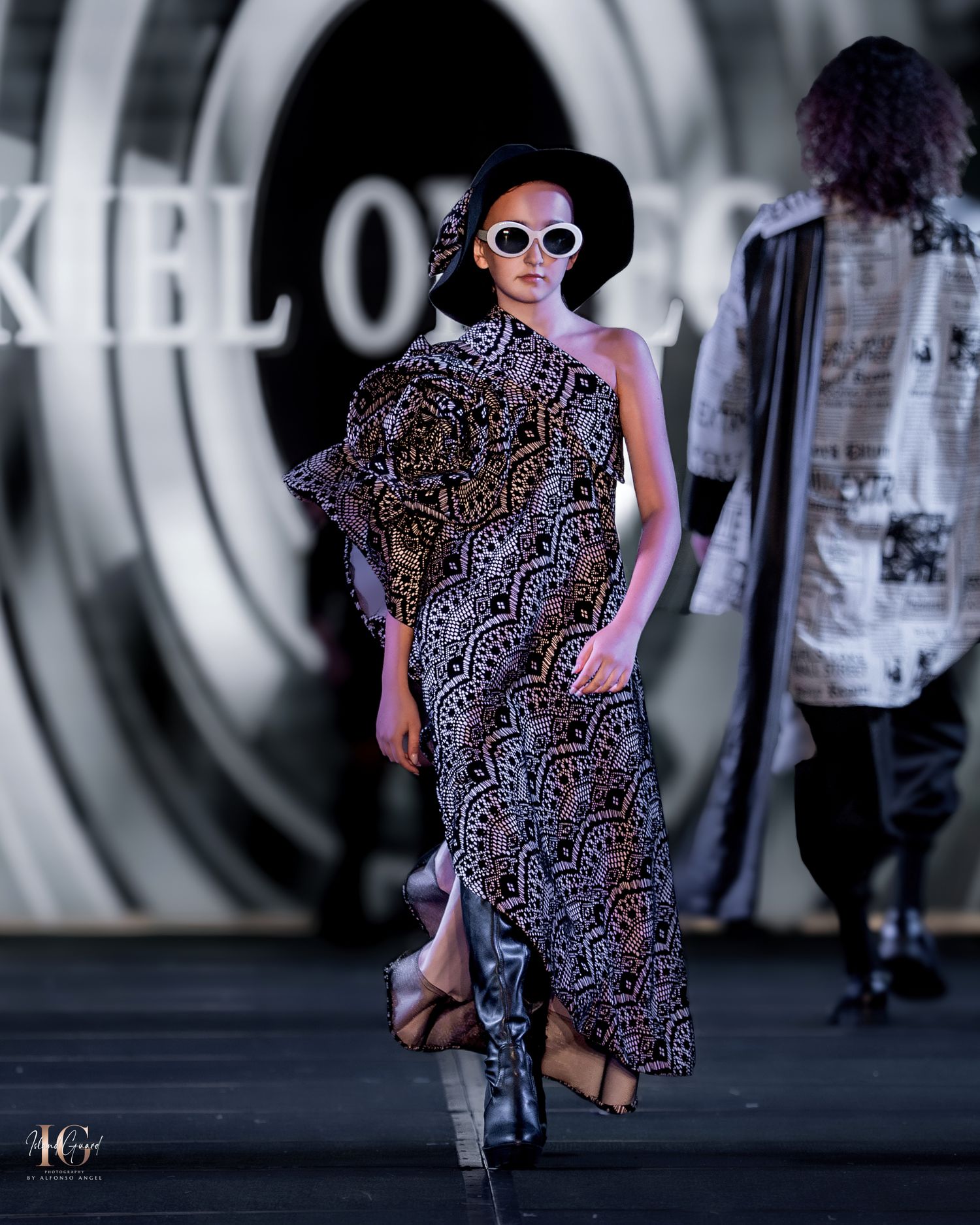 Fashion show features flowing patterned dress with black hat and sunglasses on illuminated catwalk.