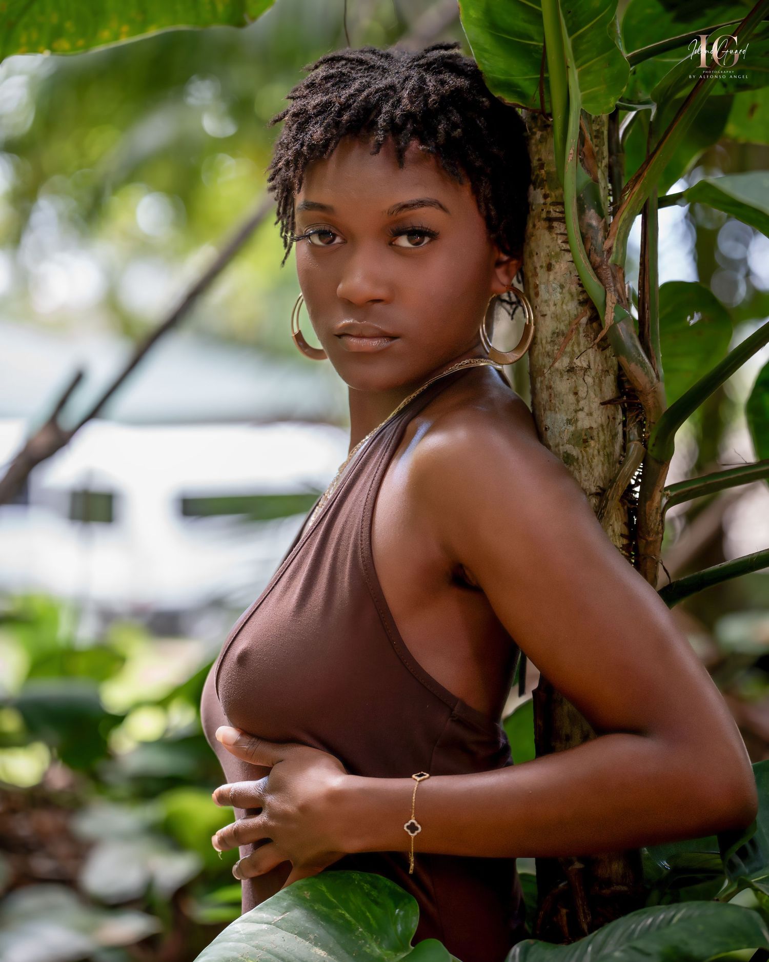 Figure in artistic photo session poses against lush green foliage in natural outdoor setting.