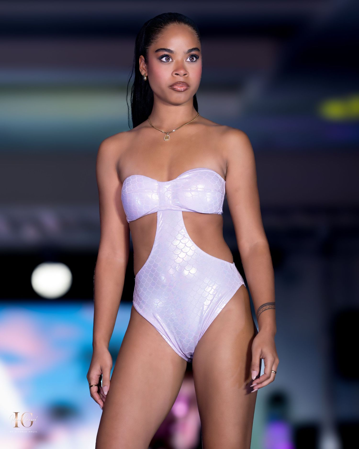 Model poses in white sequined cutout swimsuit on fashion runway with stage lighting.