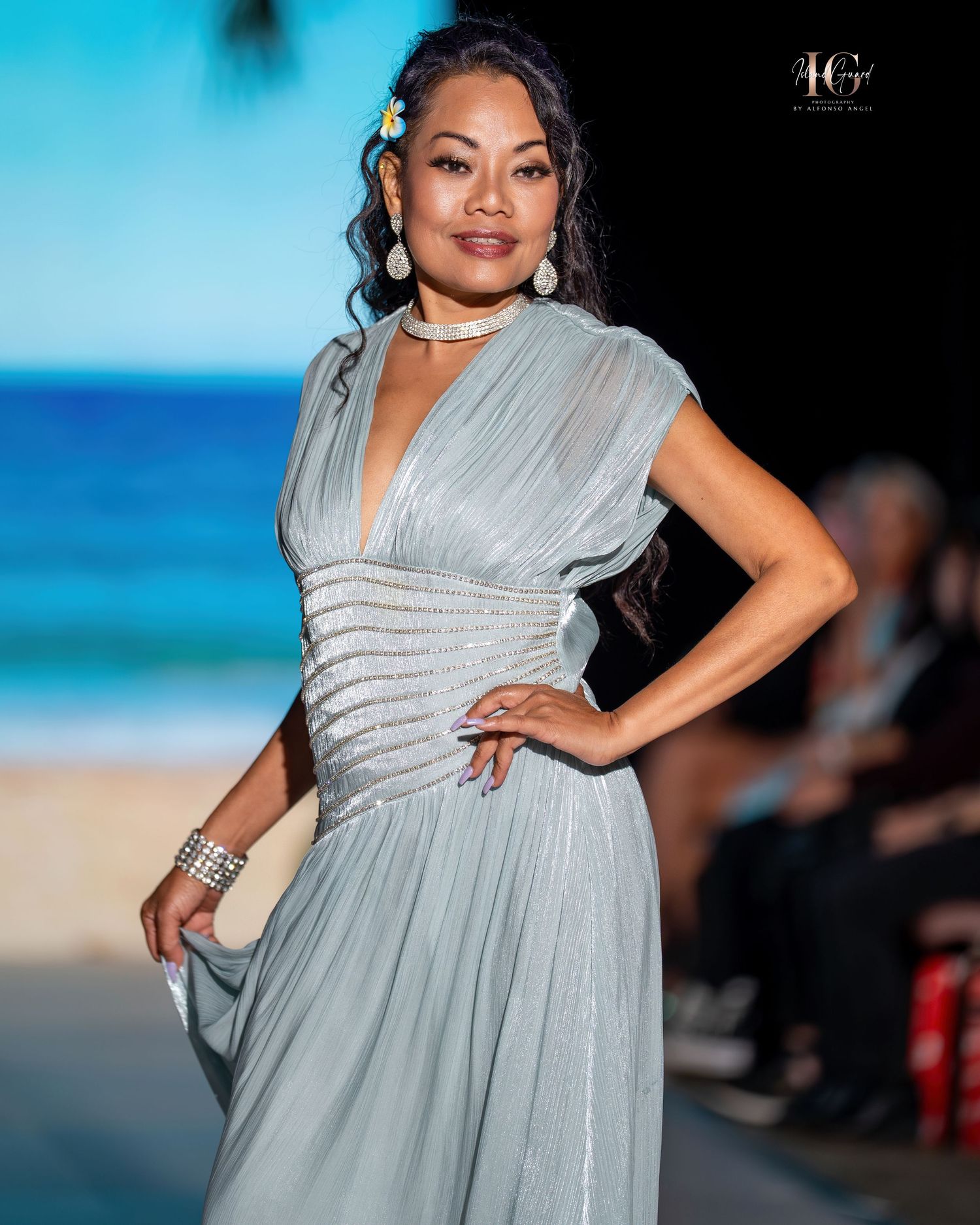 Model walks runway in flowing silver-gray evening gown with ruched waist detail at beachside fashion show.